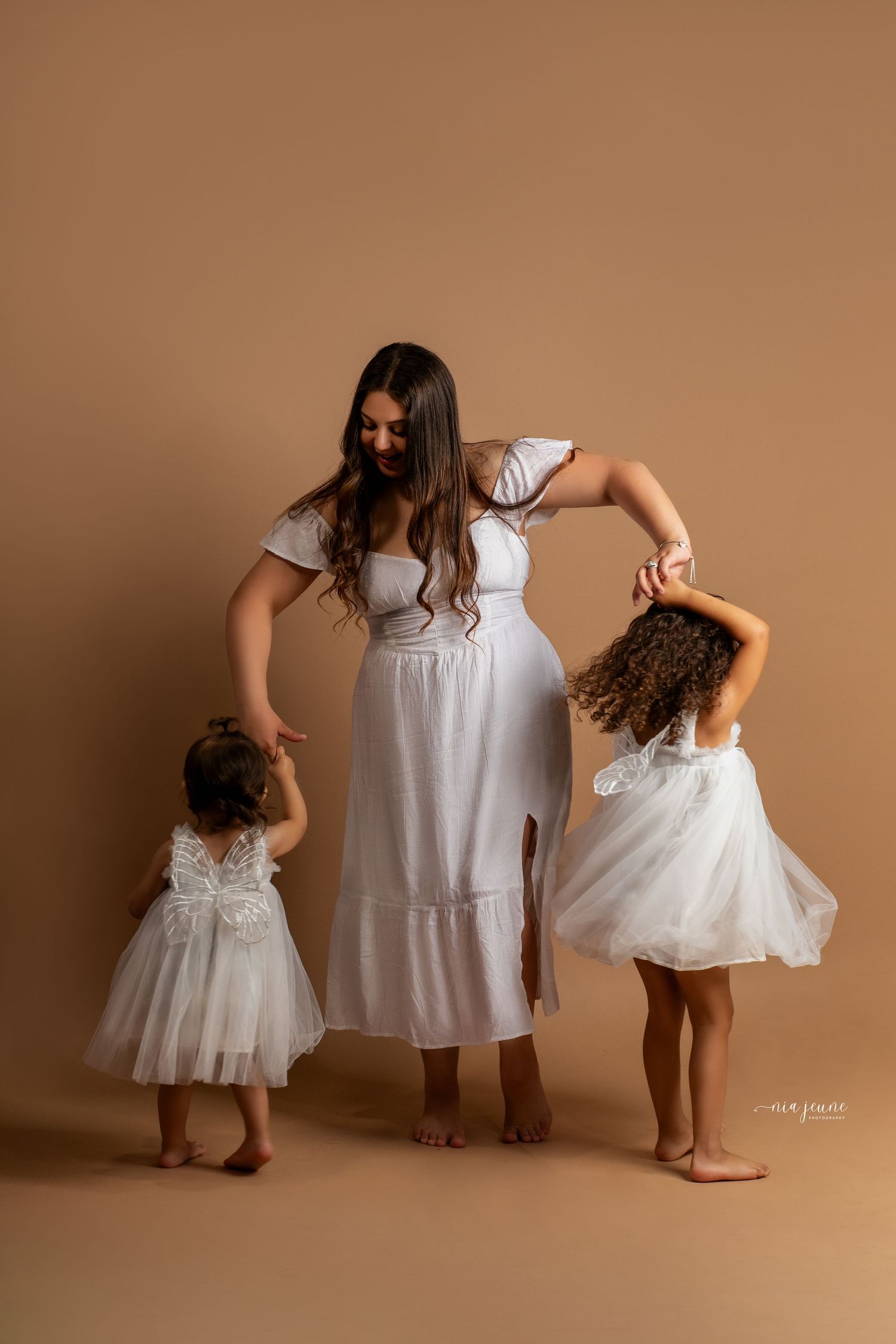 Dancing scene with mother and daughters in matching white dresses against warm brown backdrop.