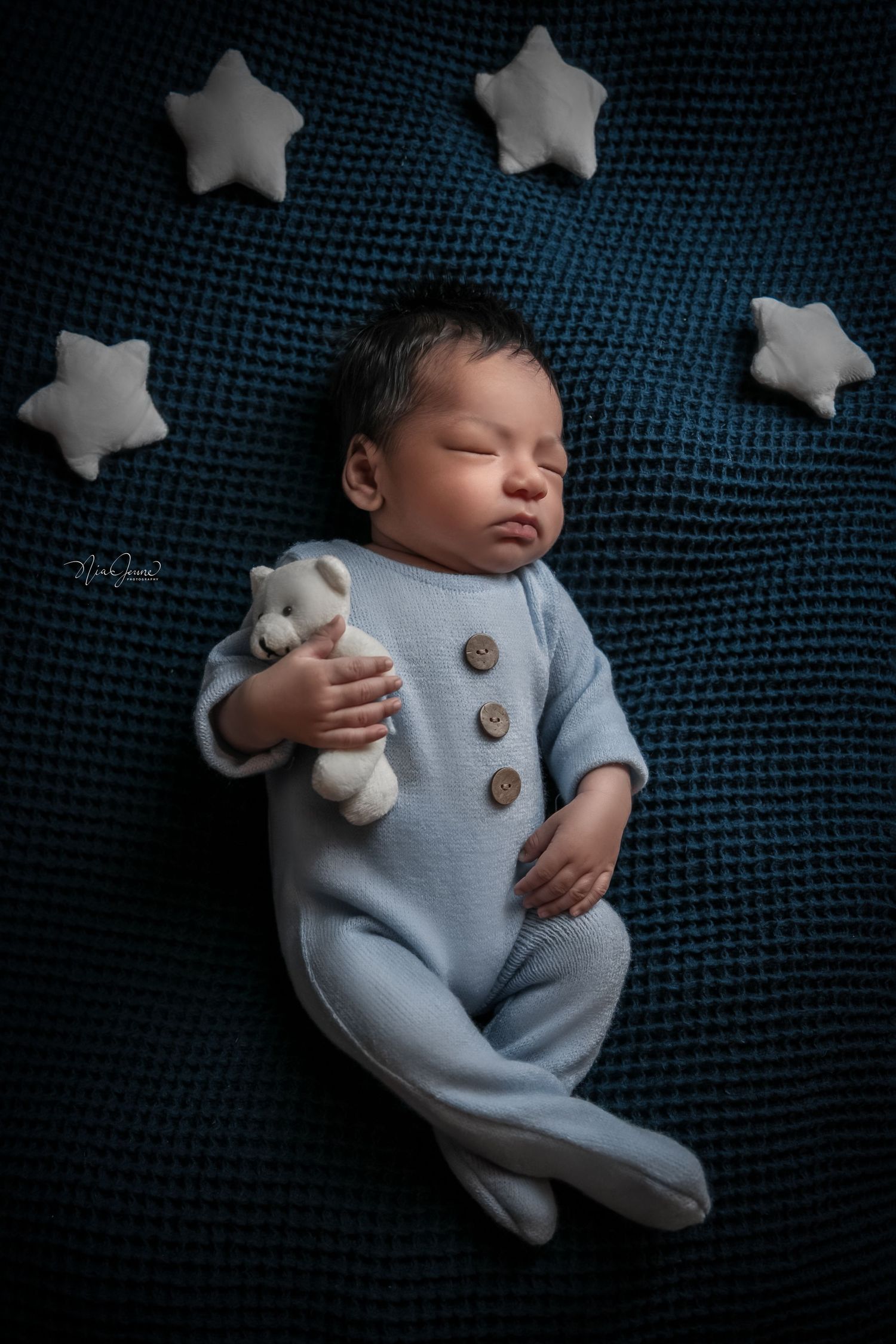 Sleeping newborn in blue onesie surrounded by decorative white stars on navy fabric.