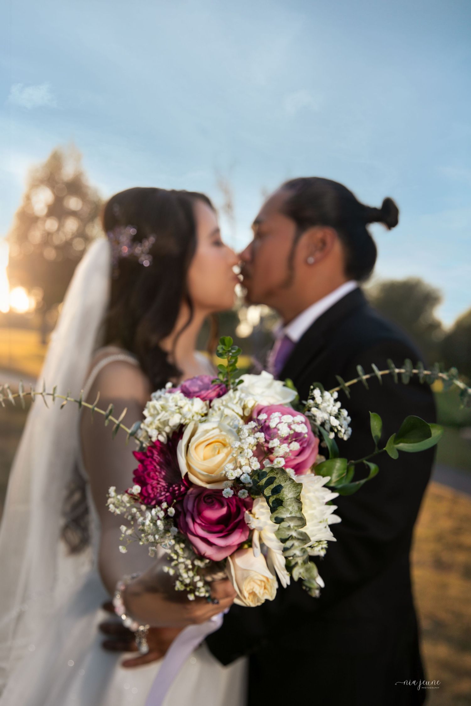 Blurred silhouettes of a bride and groom kissing at sunset with a bouquet of pink and white roses in the foreground.