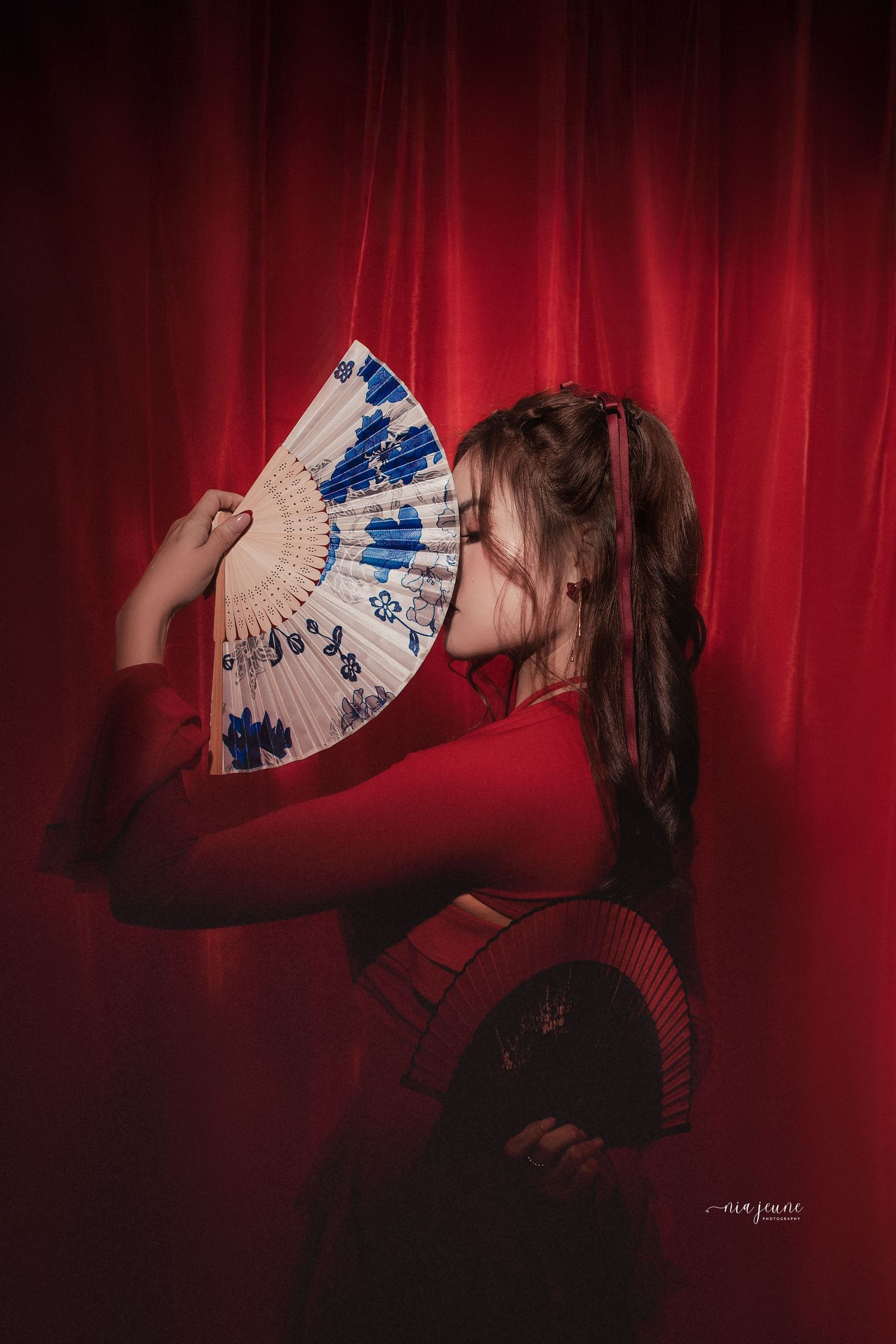 Dramatic portrait with decorative blue and white folding fan against red curtain backdrop.