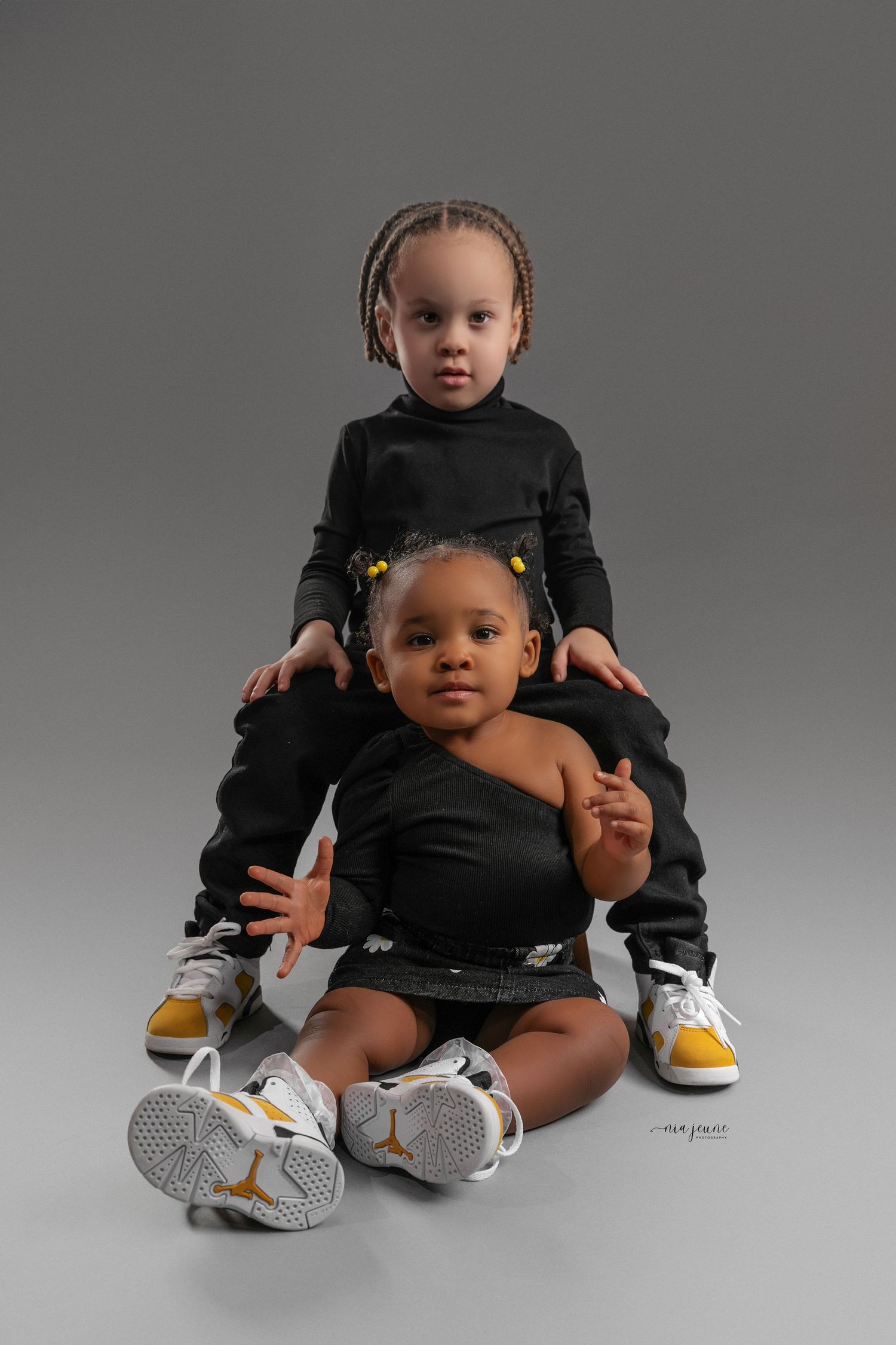 Stylish baby photoshoot featuring black outfit and yellow sneakers against grey background.