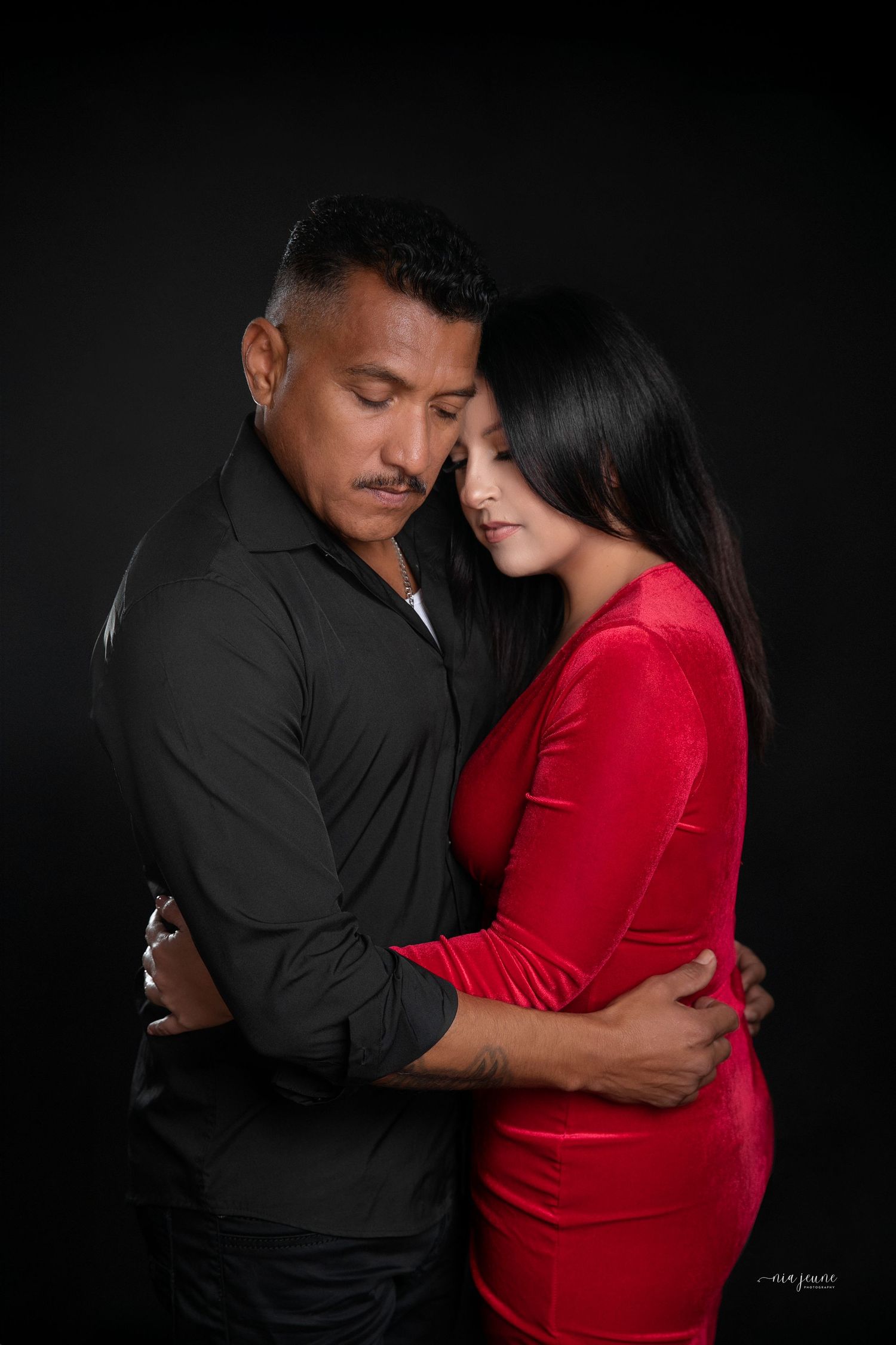 A romantic couple embracing, with woman in red dress and man in black shirt against dark background.