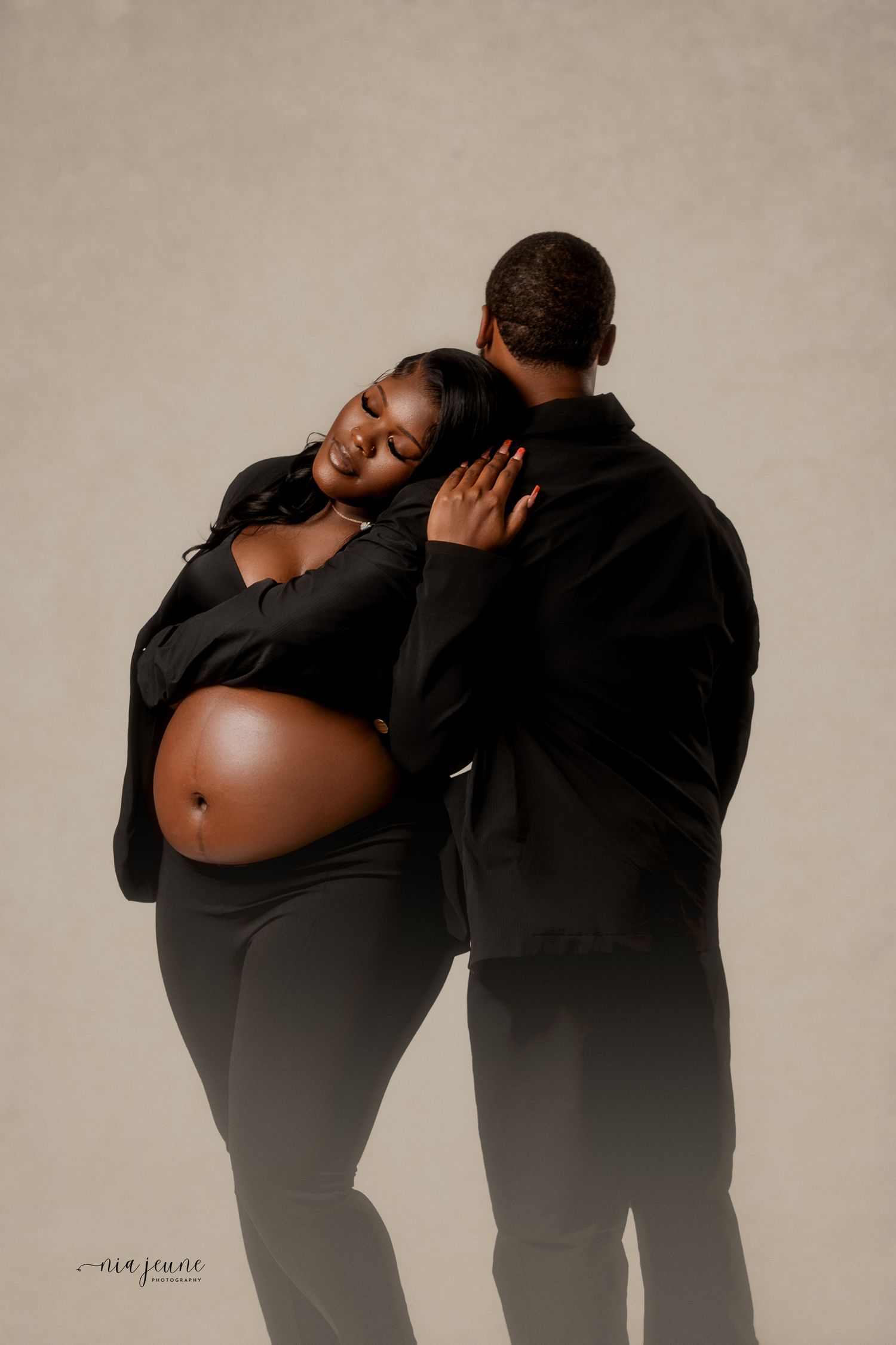 A maternity photo features an expectant couple in black clothing embracing against a neutral backdrop in a studio setting.