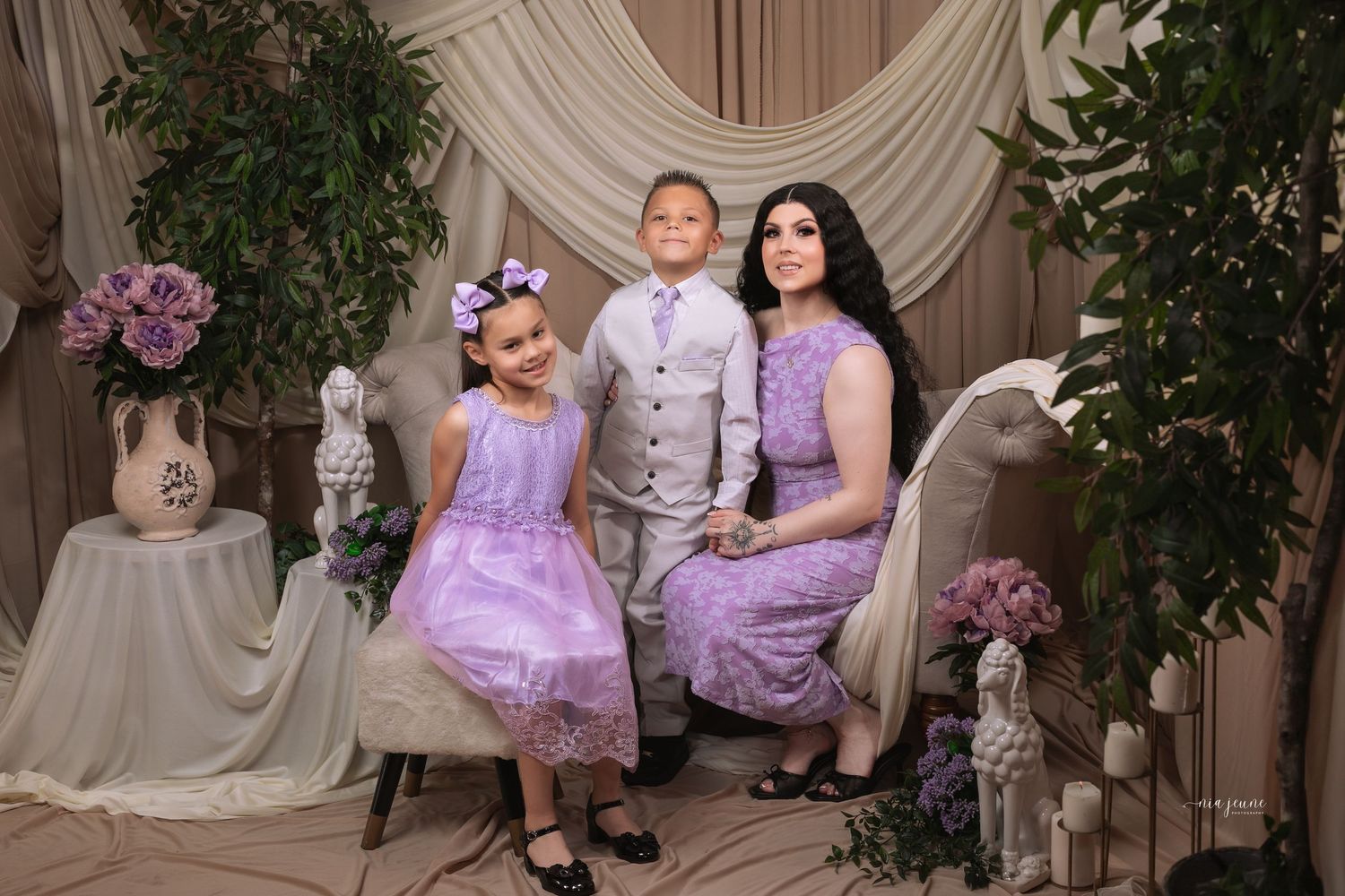 A woman in lavender lace dress poses with two children in matching purple outfits in an elegant floral studio setting.