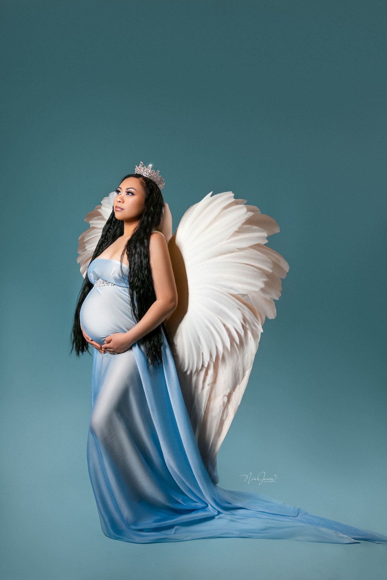A maternity photo shoot featuring a woman in blue fabric with white angel wings against a teal backdrop.