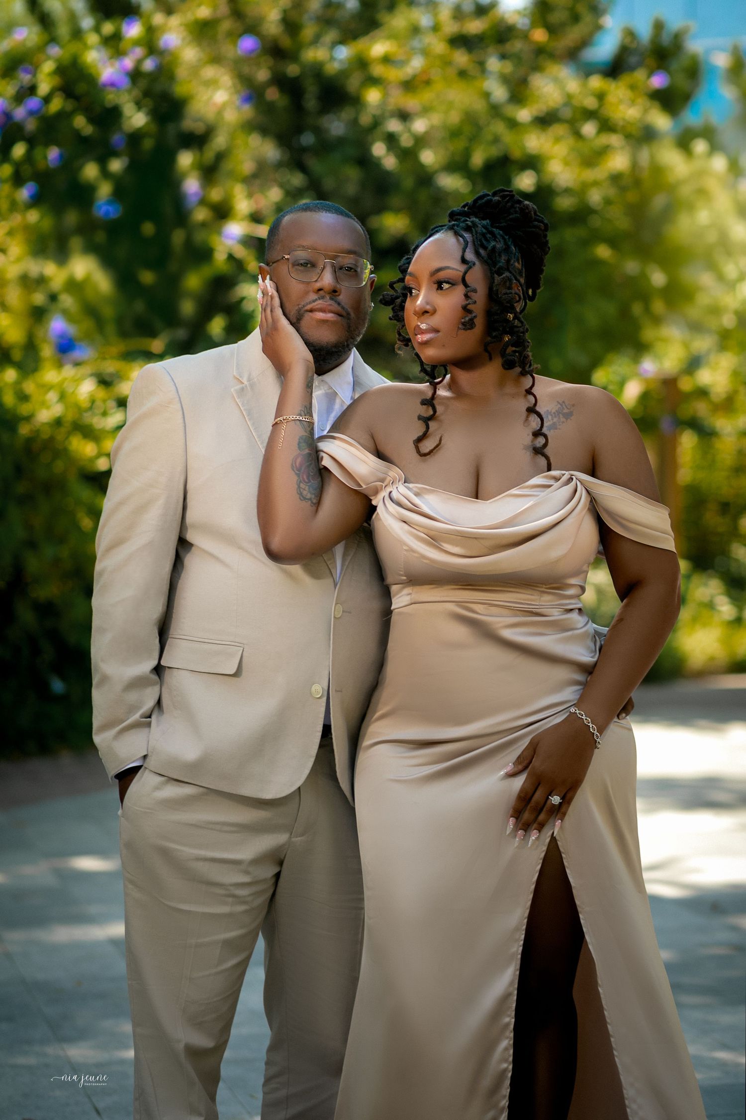 A couple poses together outdoors wearing matching beige formal attire on a sunny day with trees in the background.