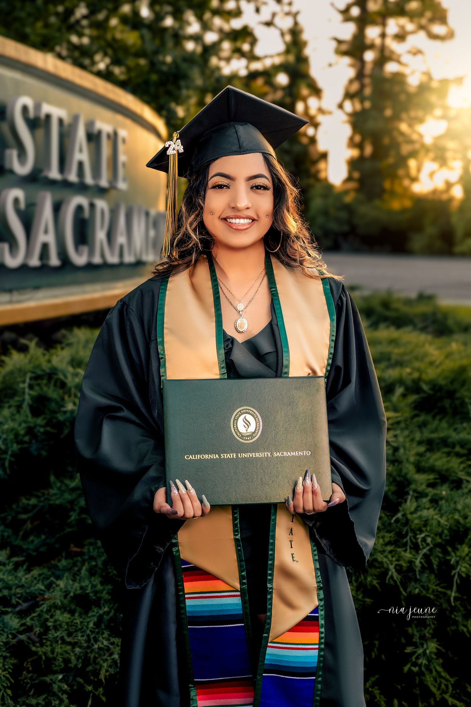 Graduate in cap and gown holding diploma at Sacramento State during golden hour.