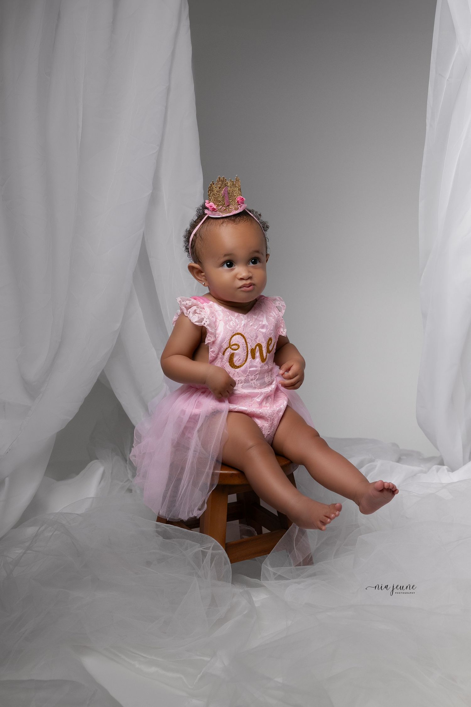 A baby in a pink tulle dress and tiara sits on a wooden stool against white fabric.