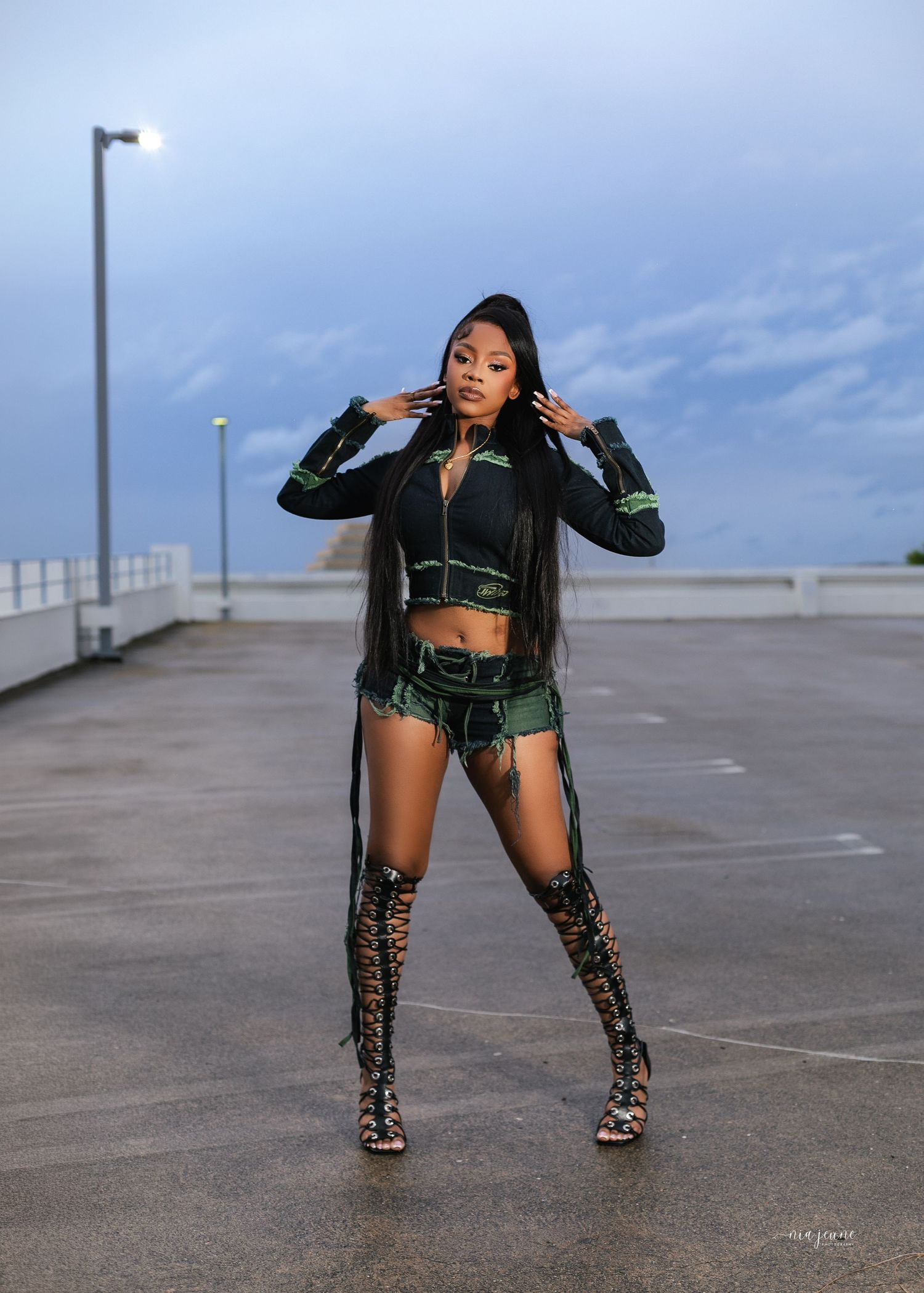 Woman in green and black outfit with thigh-high boots poses confidently on rooftop parking lot at dusk.