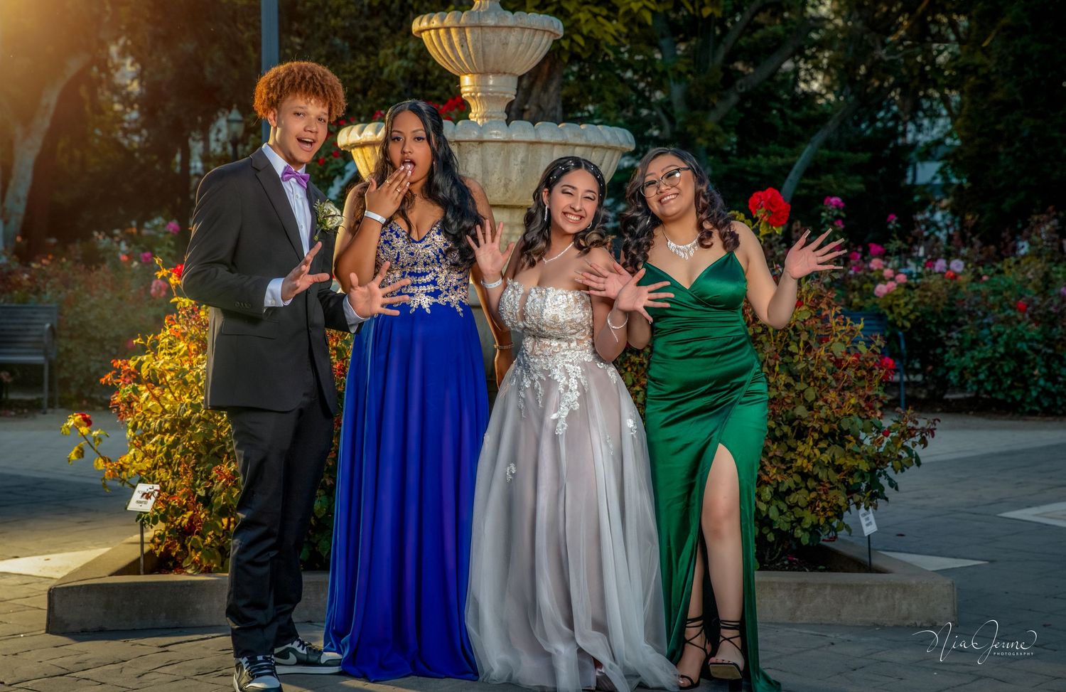 Formal group poses outdoors at evening event near illuminated fountain and garden area.