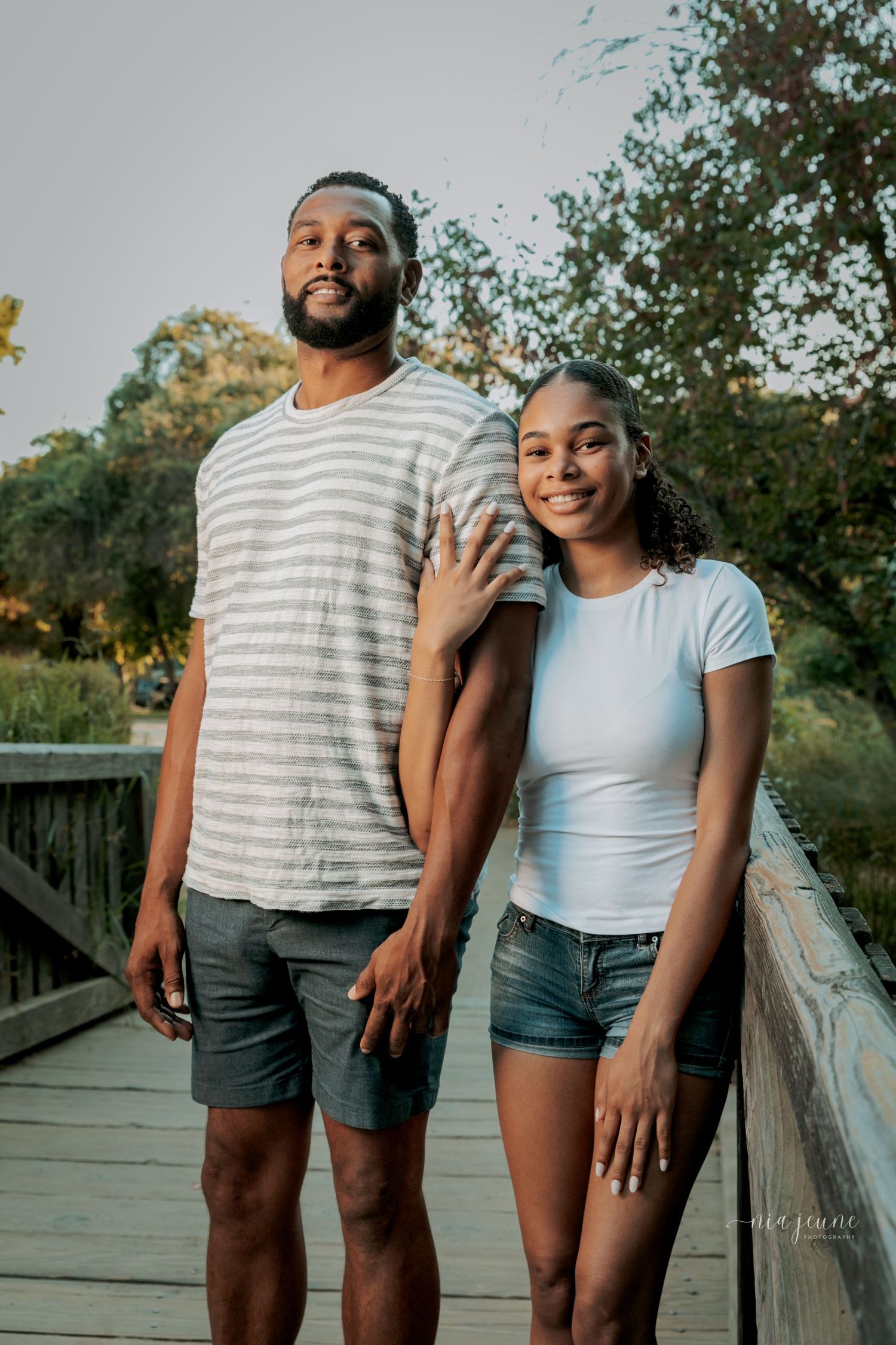 Couple in casual summer outfits poses together on wooden bridge during sunset.