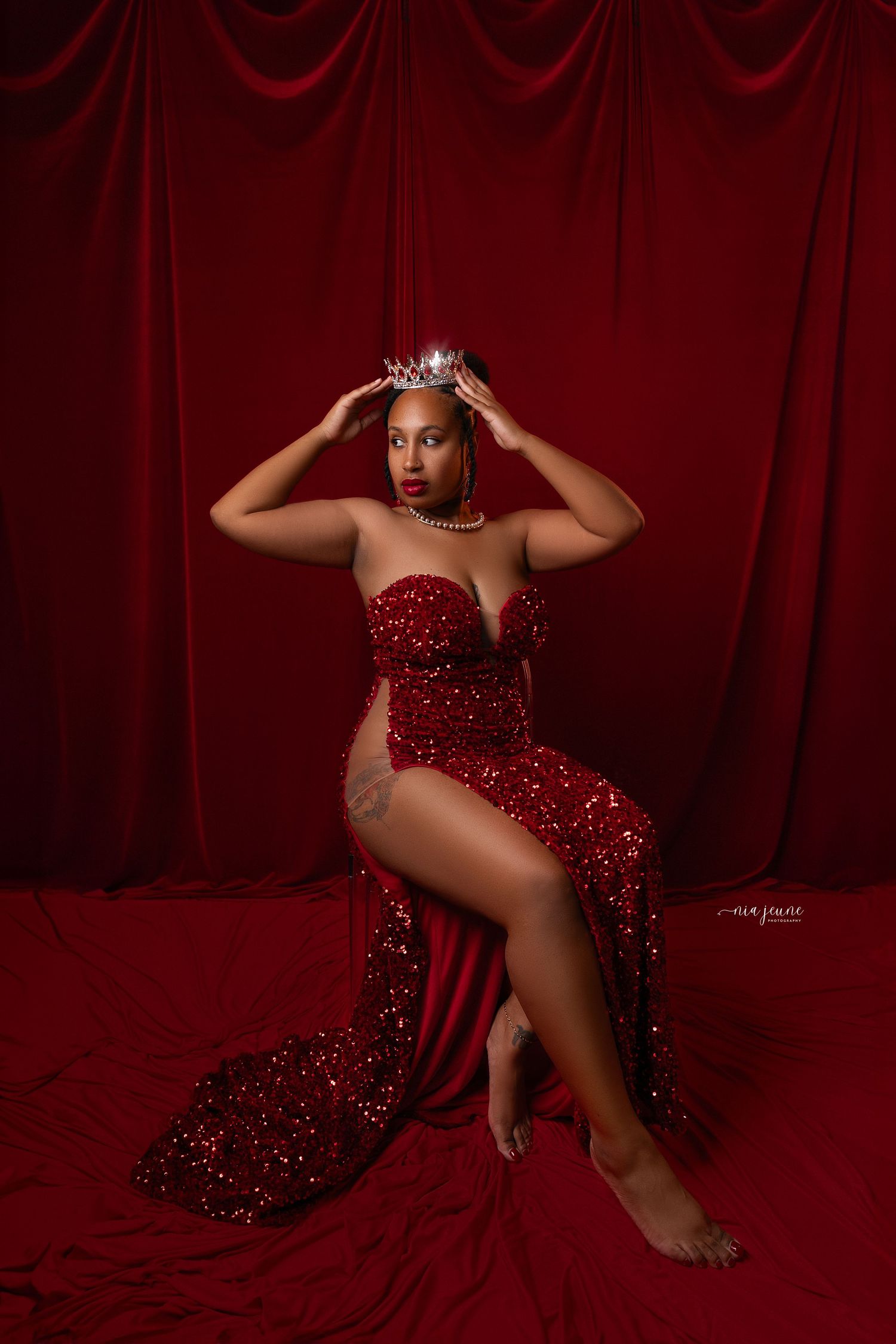 Glamorous pose in sparkling red gown and crown against deep red velvet backdrop.