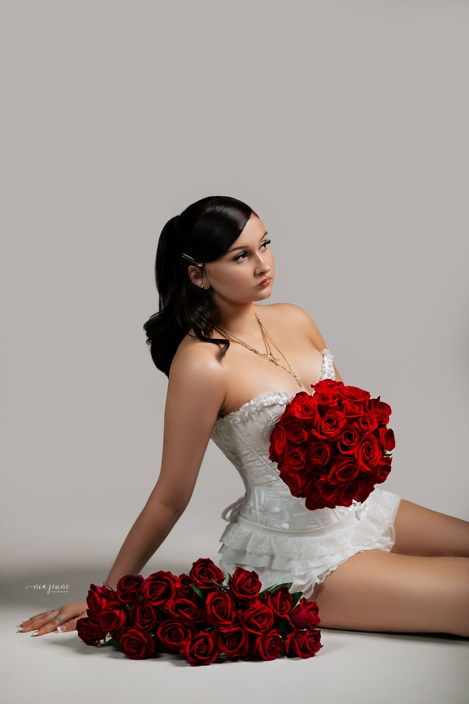 Elegant figure in white lace corset dress posing with vibrant red roses against gray backdrop in studio setting.