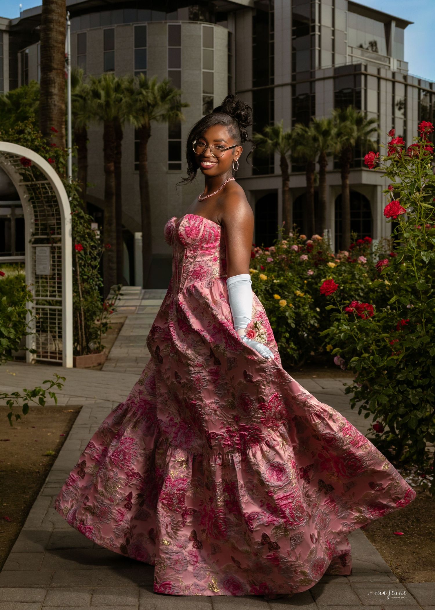 Young woman in a pink floral strapless ball gown with white gloves poses outdoors near roses.