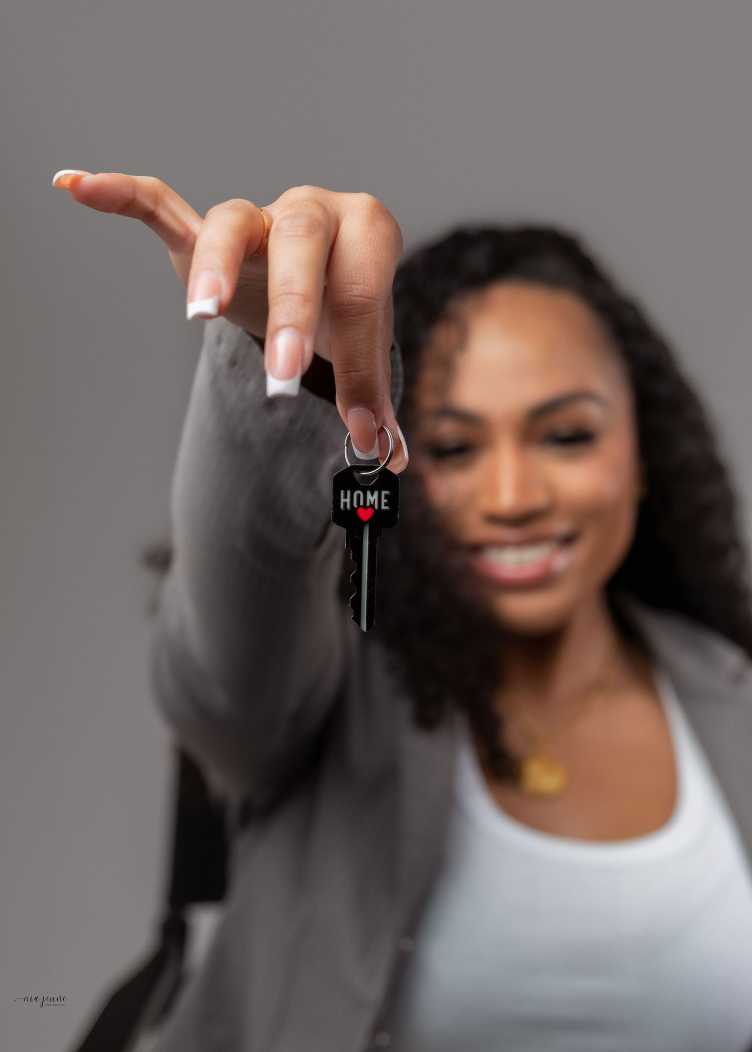 Smiling woman holding a house key with a red HOME keychain toward the camera against a gray background.