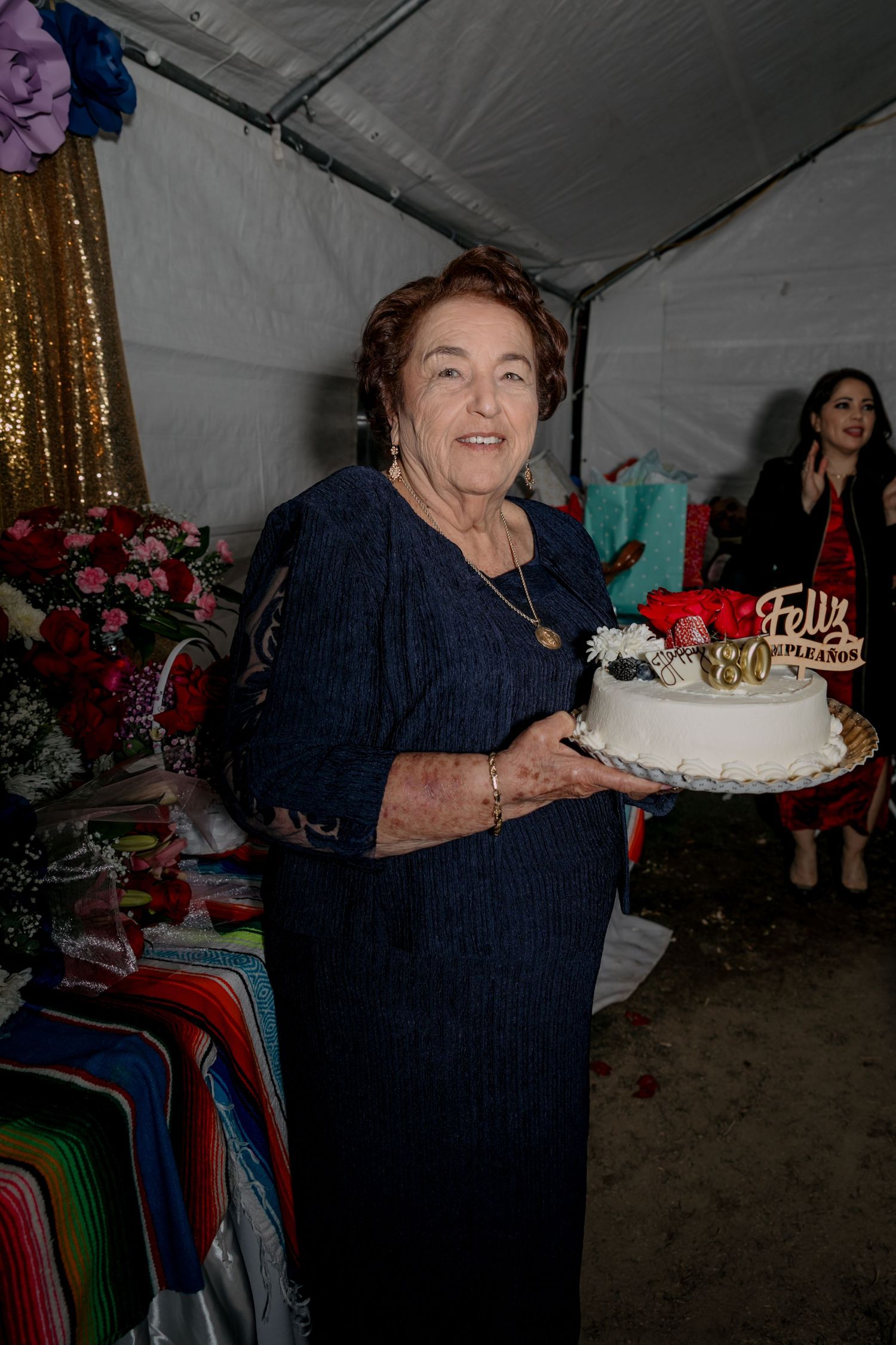 Woman in navy dress holds decorated birthday cake at celebration under white tent.