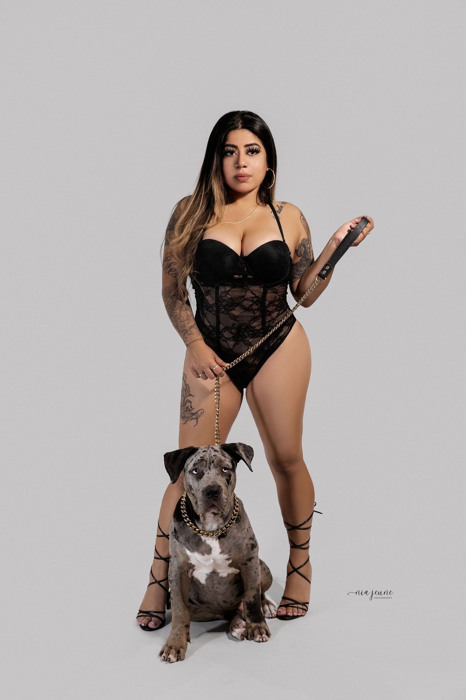 A woman in black lingerie poses with a grey pit bull dog against a studio backdrop.