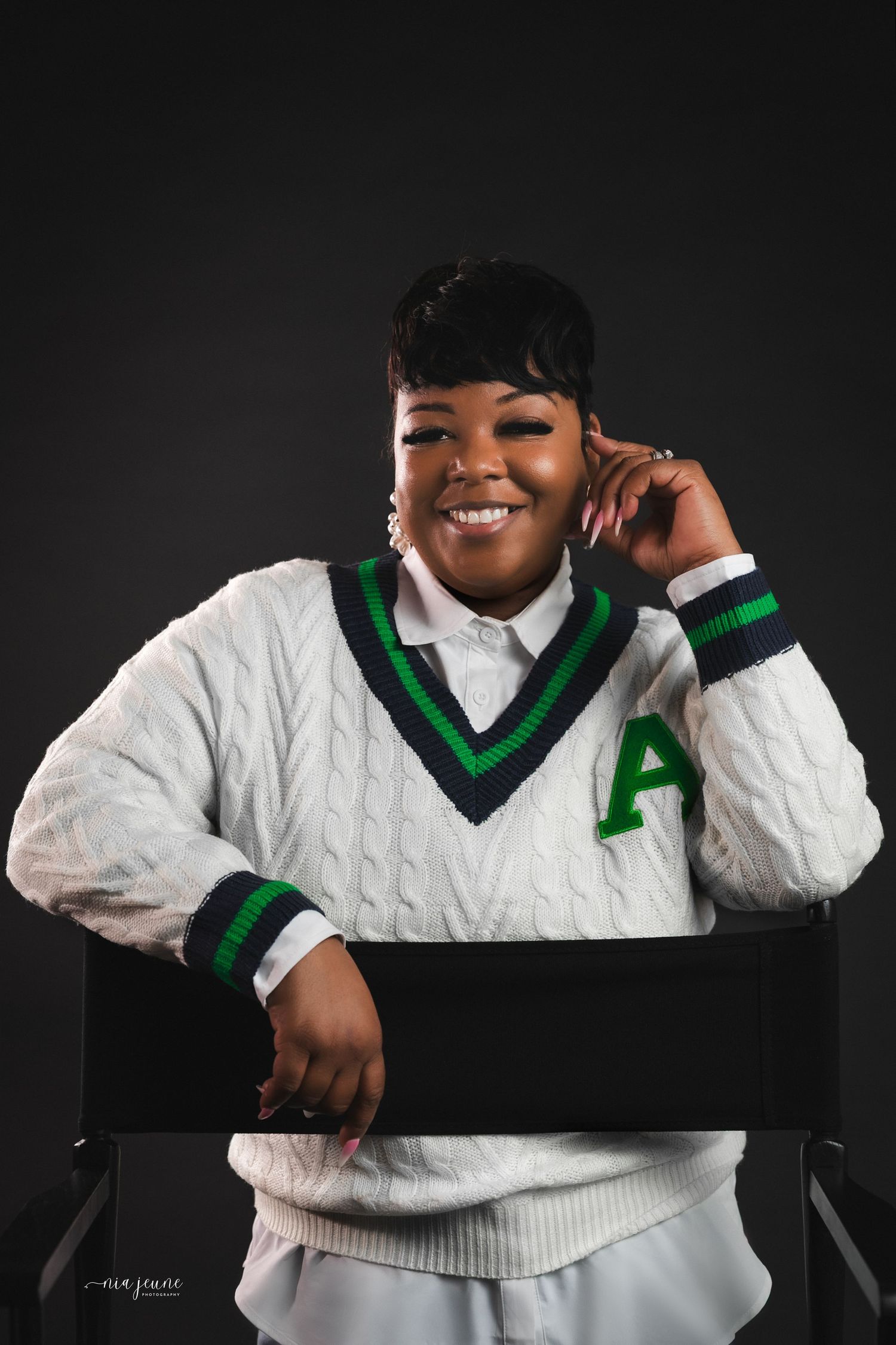 A person wearing a white cable-knit varsity sweater with green and black trim poses against a dark background.