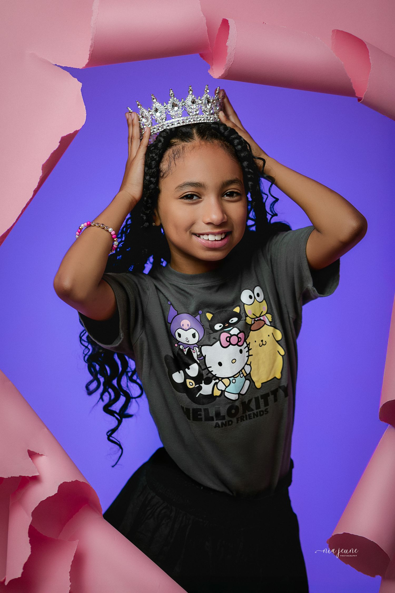 Young person wearing a Hello Kitty t-shirt and sparkly crown poses against purple background with pink torn paper frame.