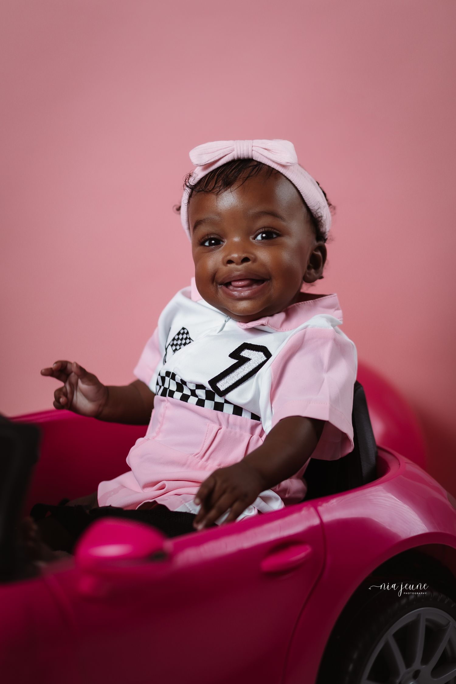 Baby wearing pink outfit and white headband sits in pink toy car against rose-colored background.