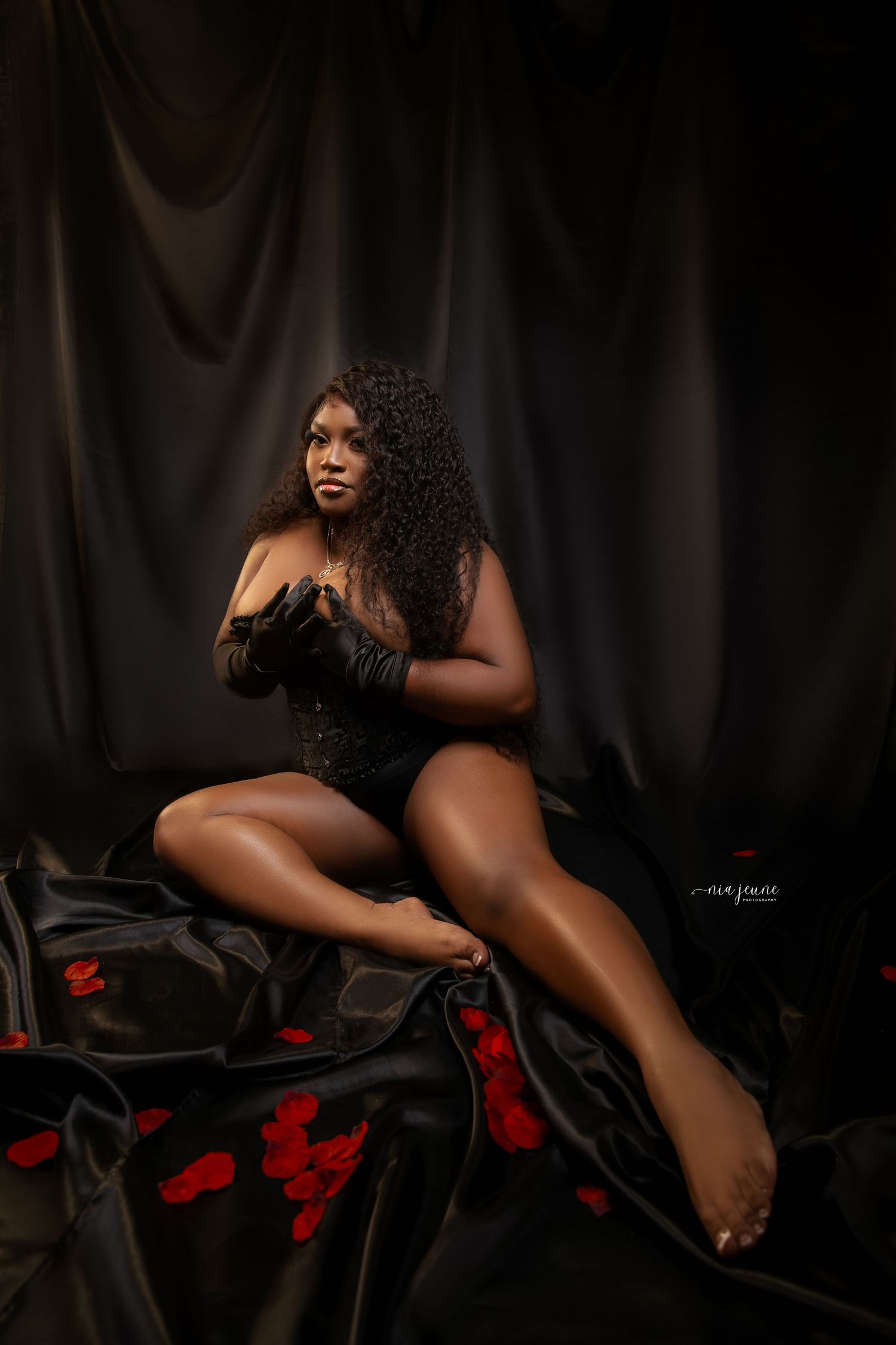 Boudoir style portrait in black lingerie on dark fabric with red rose petals.