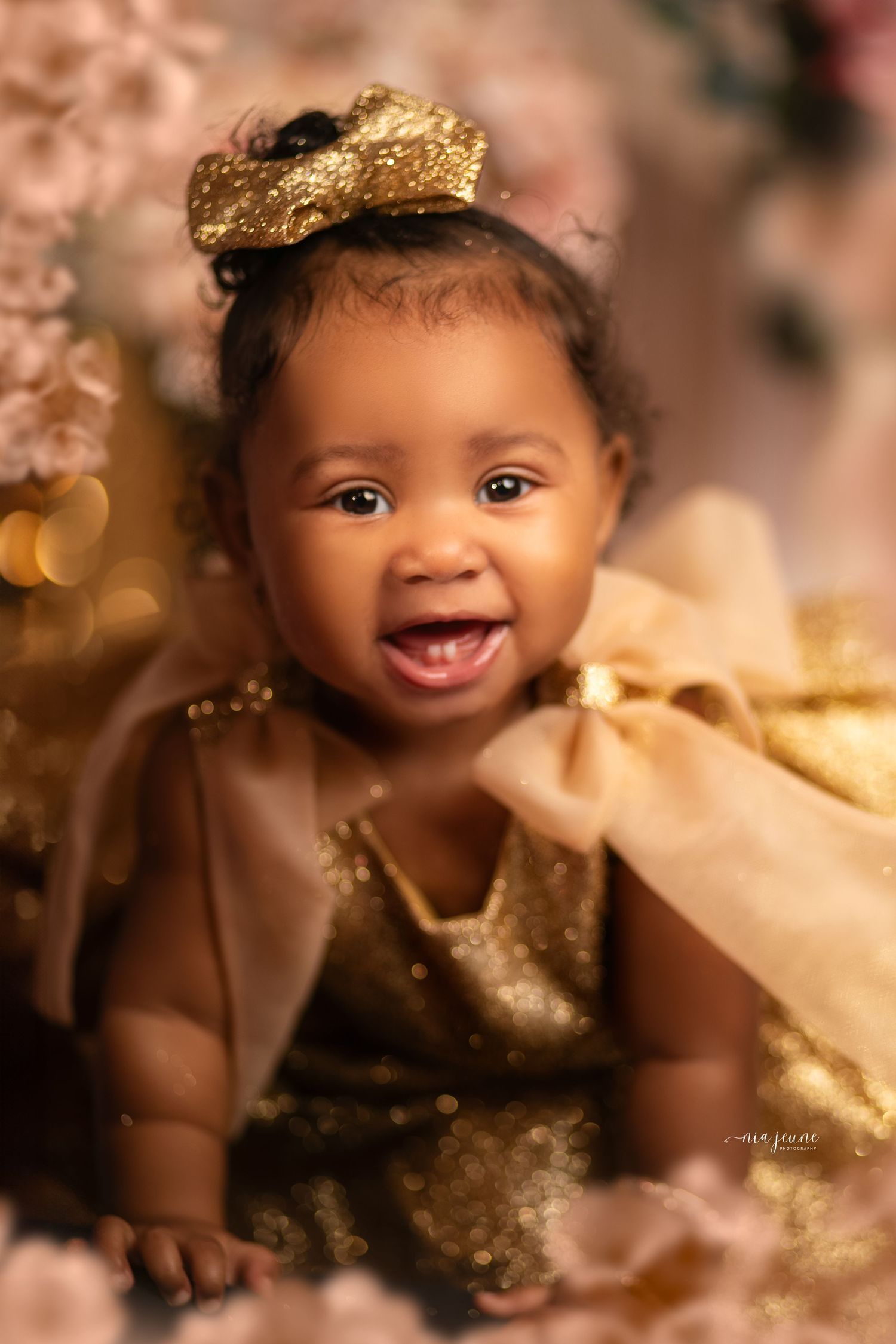 Baby wearing gold crown and dress smiles brightly amid pink and gold themed setting.