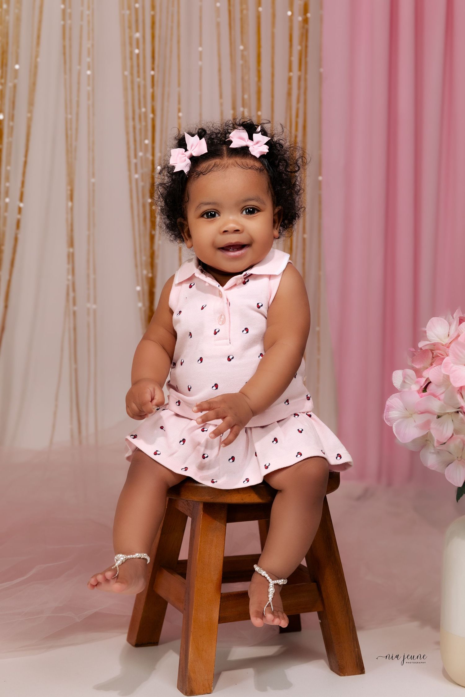 A happy baby in a pink dress with small floral details sits on a wooden stool against a pink and gold backdrop.