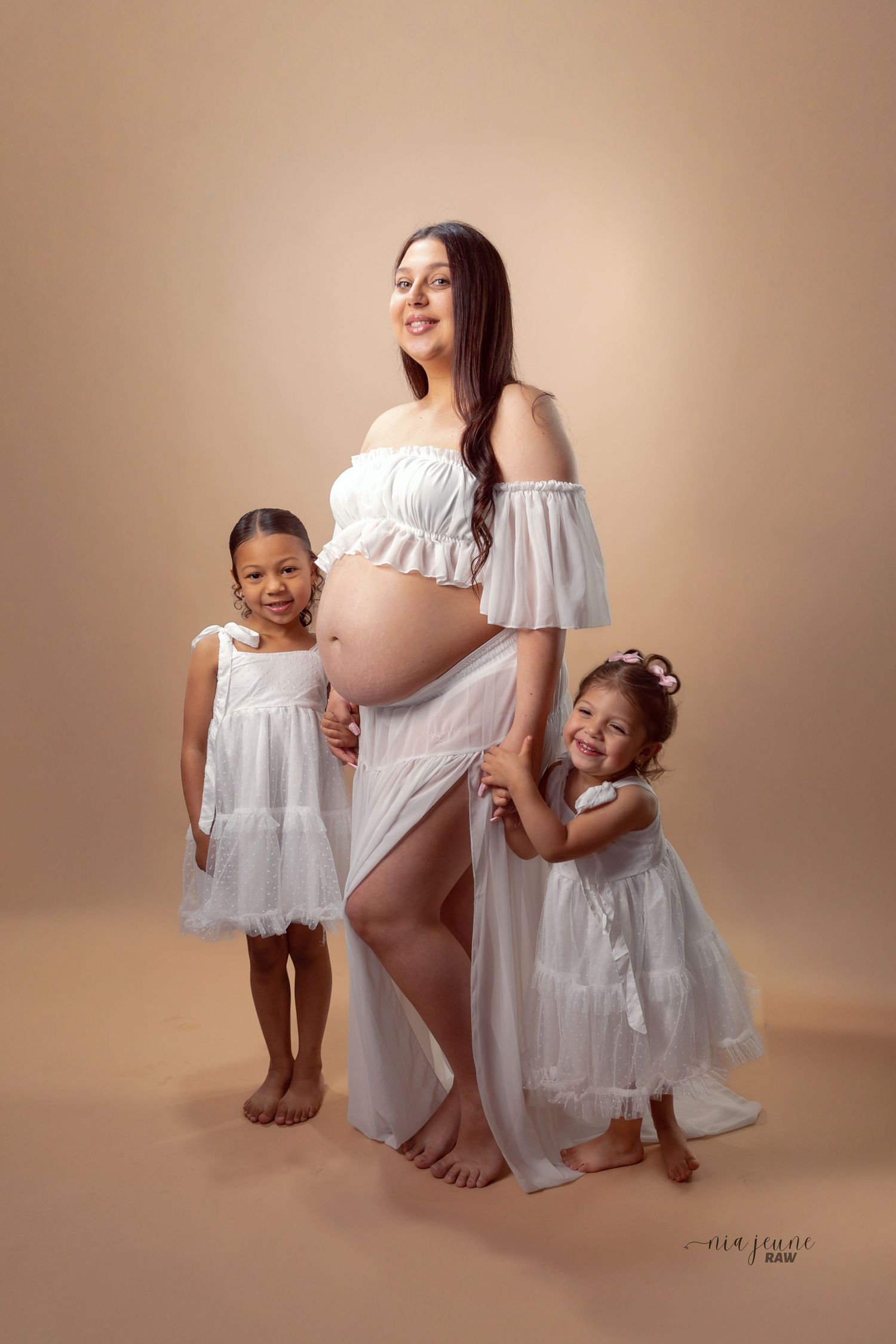 A maternity portrait with two young children all wearing white dresses against a neutral backdrop.