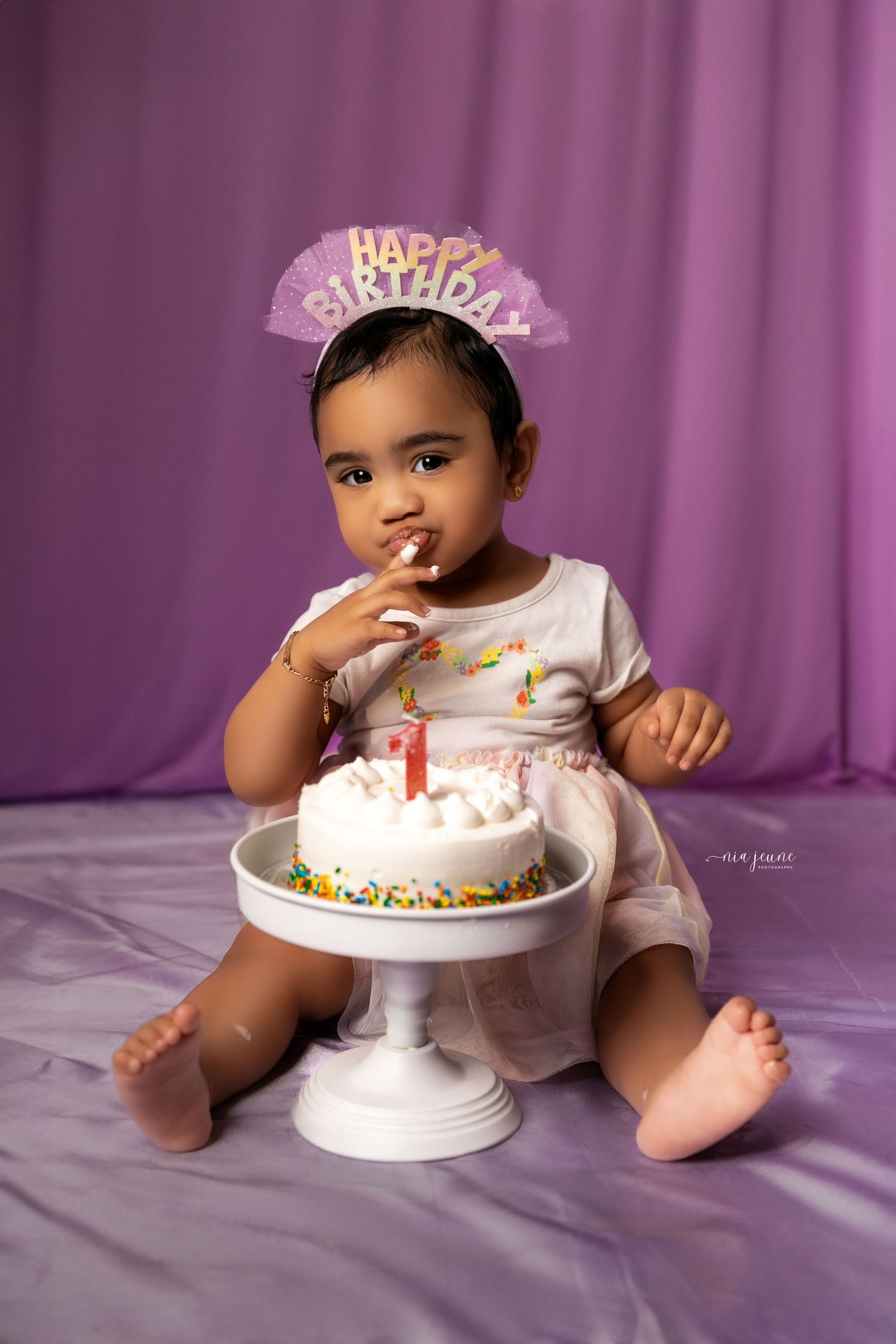 Baby in white dress celebrating first birthday with cake and pink backdrop.