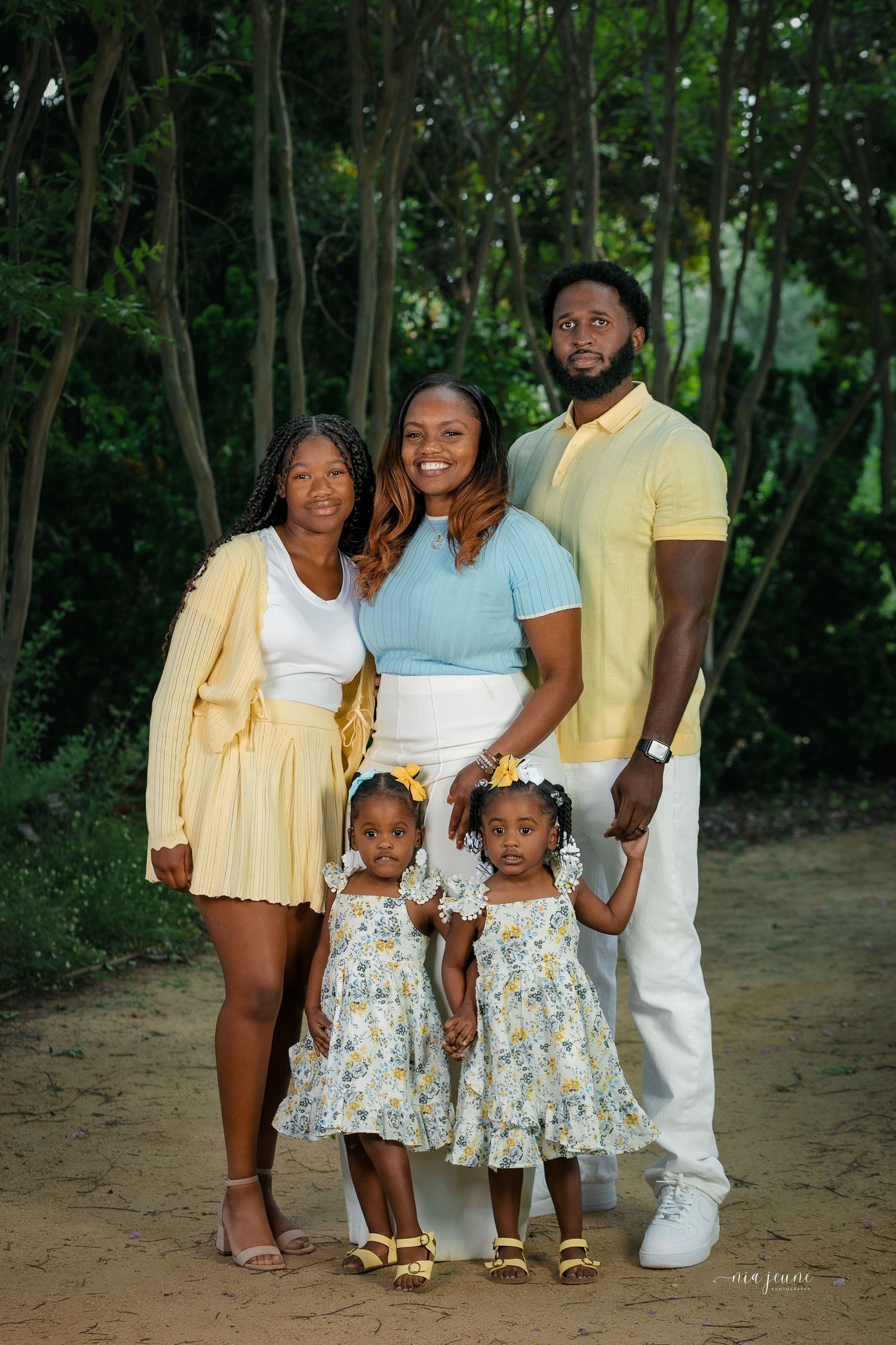 Family portrait with matching outfits and twin girls in floral dresses against wooded background.