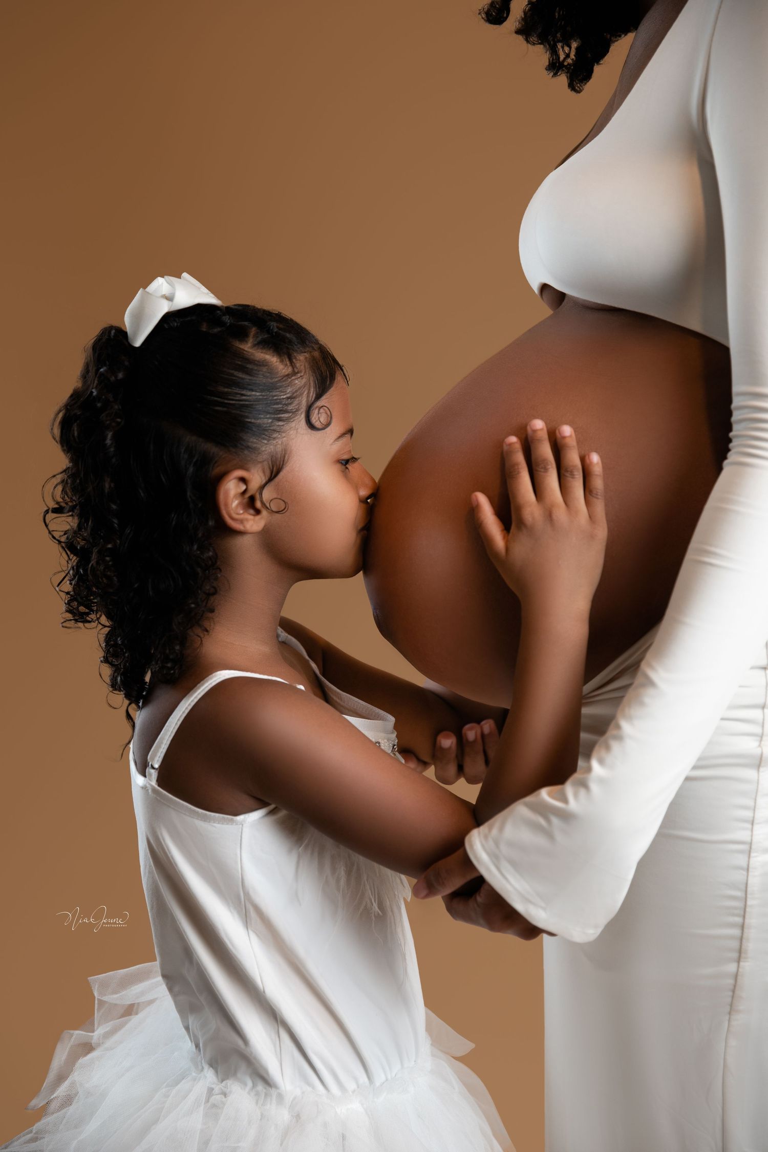 Sweet moment between child and expectant mother in white dress against brown background.