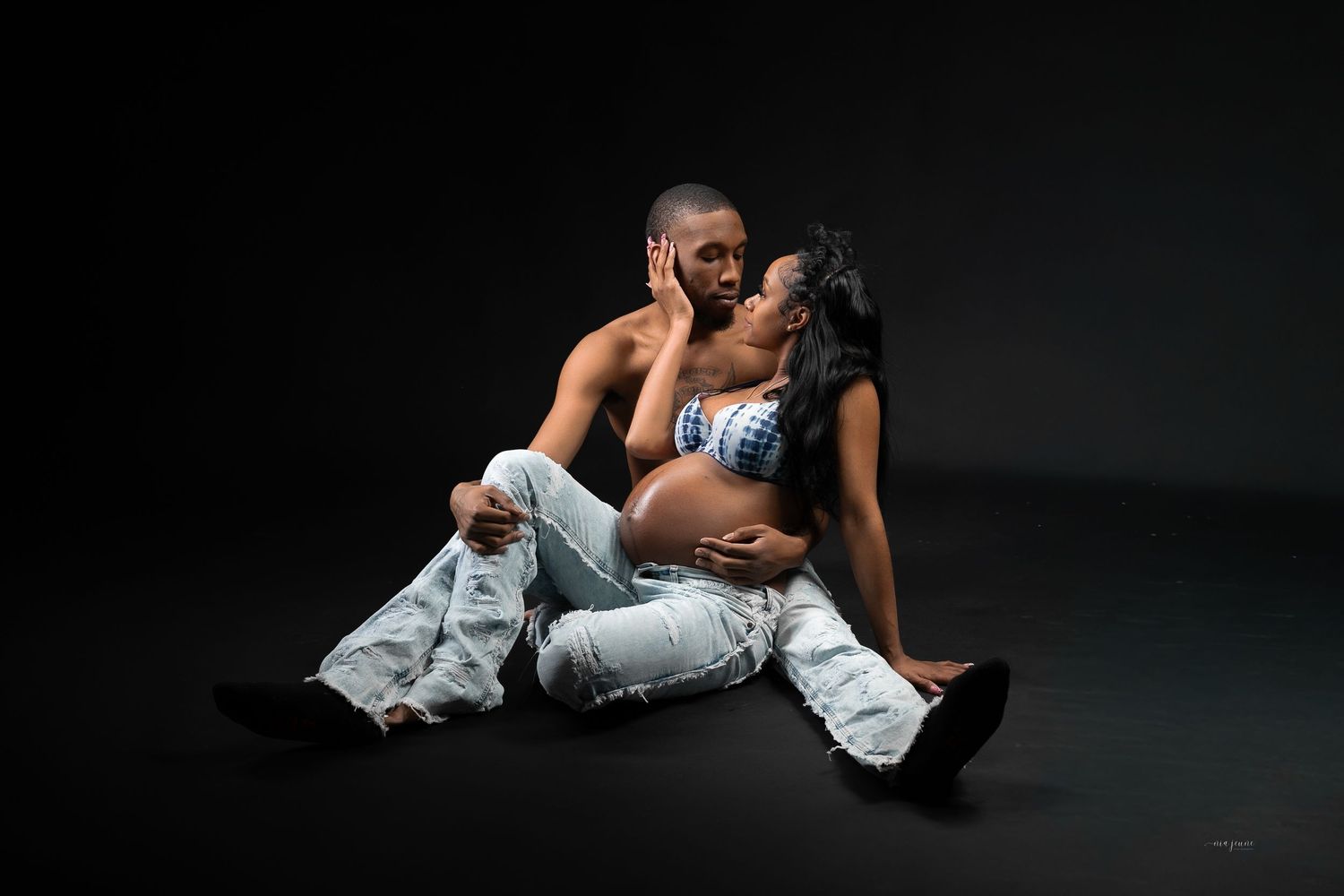 Couple sharing intimate moment, pregnant woman in ripped jeans with shirtless partner on dark studio floor.