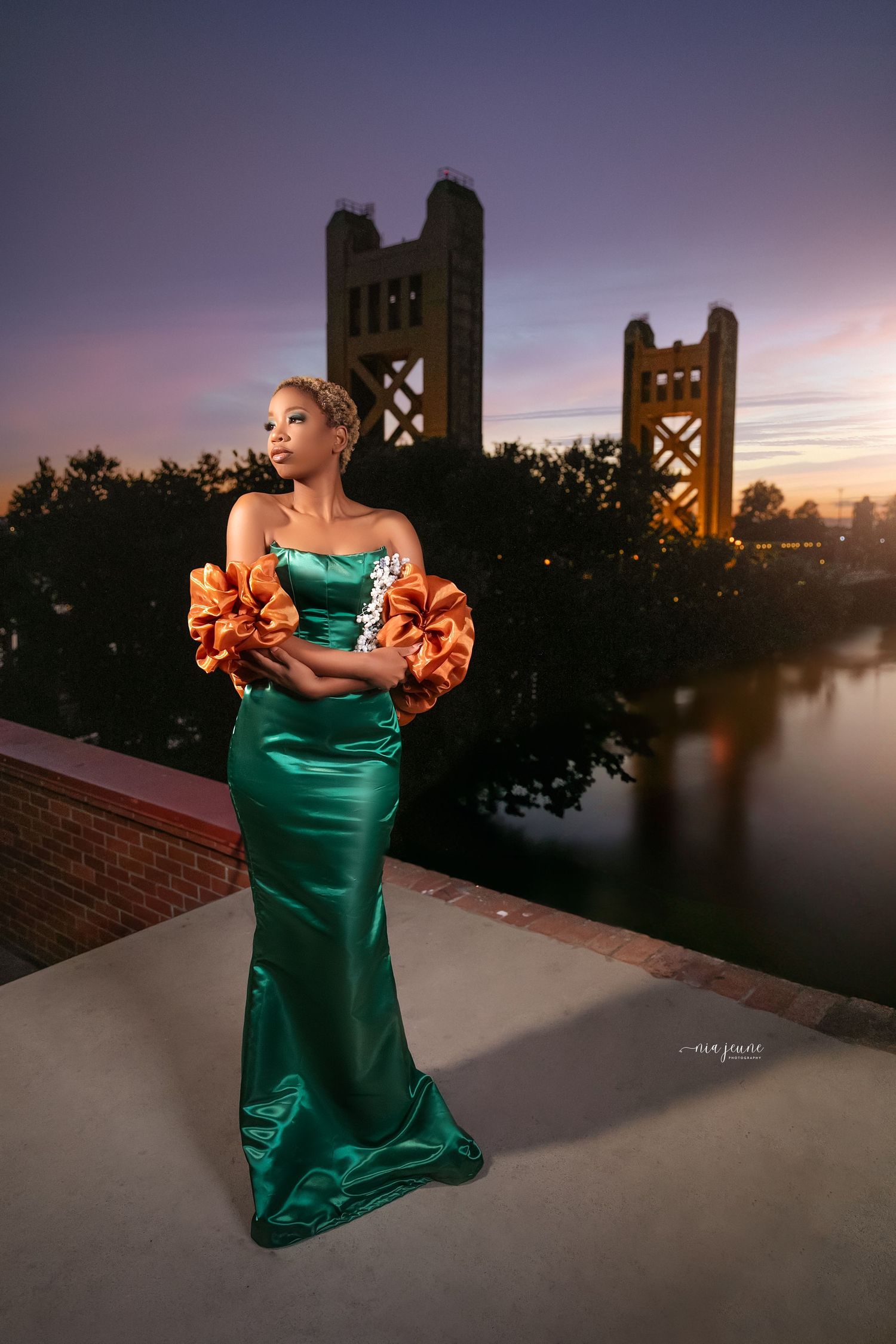 Evening portrait in emerald gown with orange shawl against Sacramento Tower Bridge at sunset.