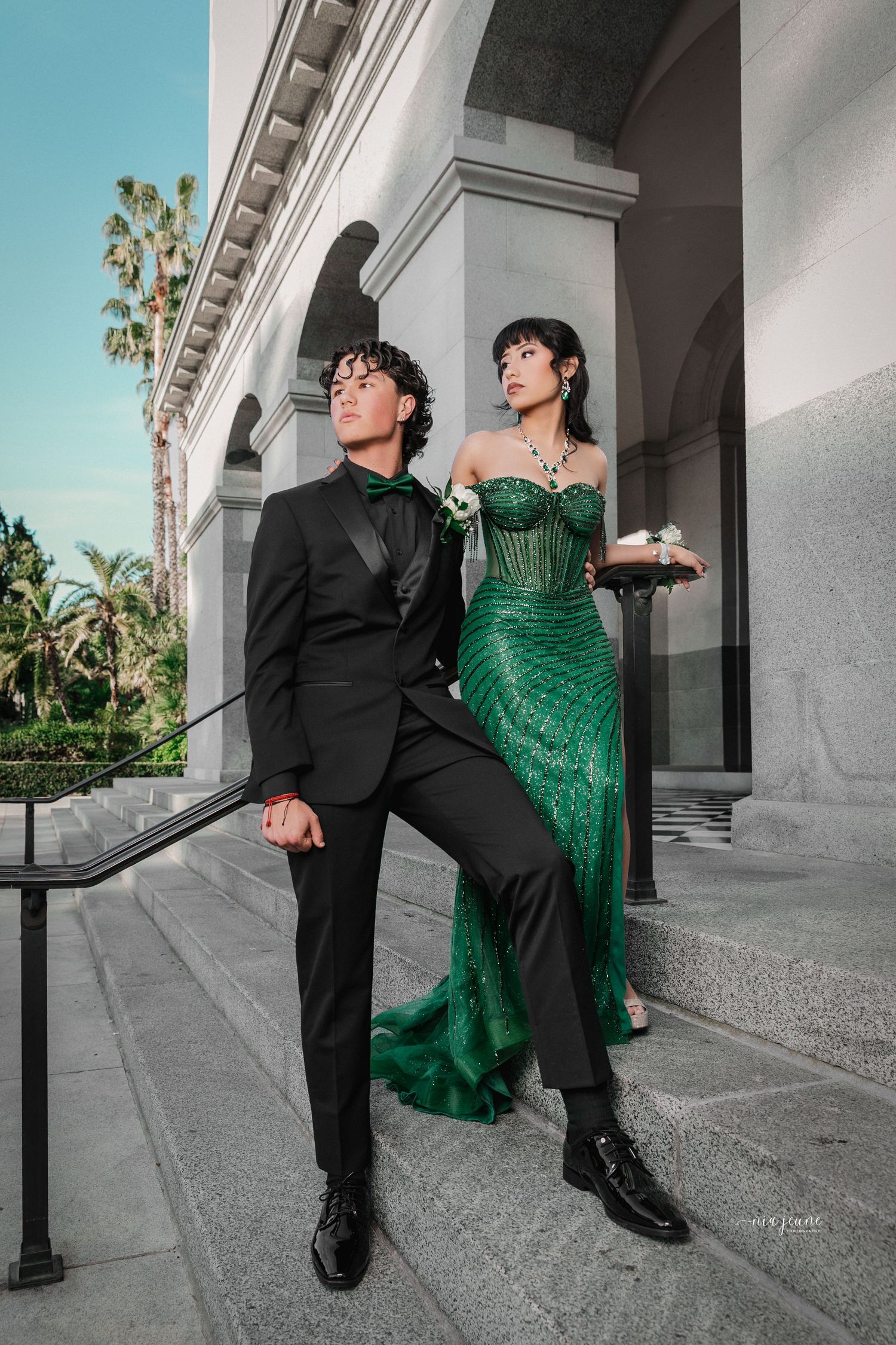 A couple poses dramatically in formal wear, with a black suit and emerald green gown against classical architecture.