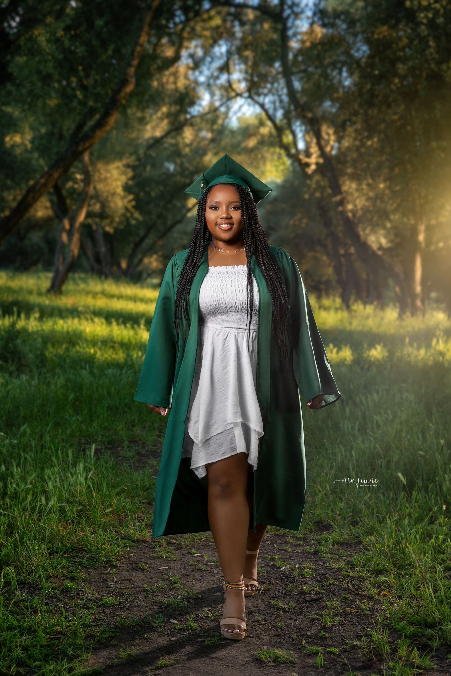 Graduate in emerald cap and gown poses on nature path with golden sunlight filtering through trees.