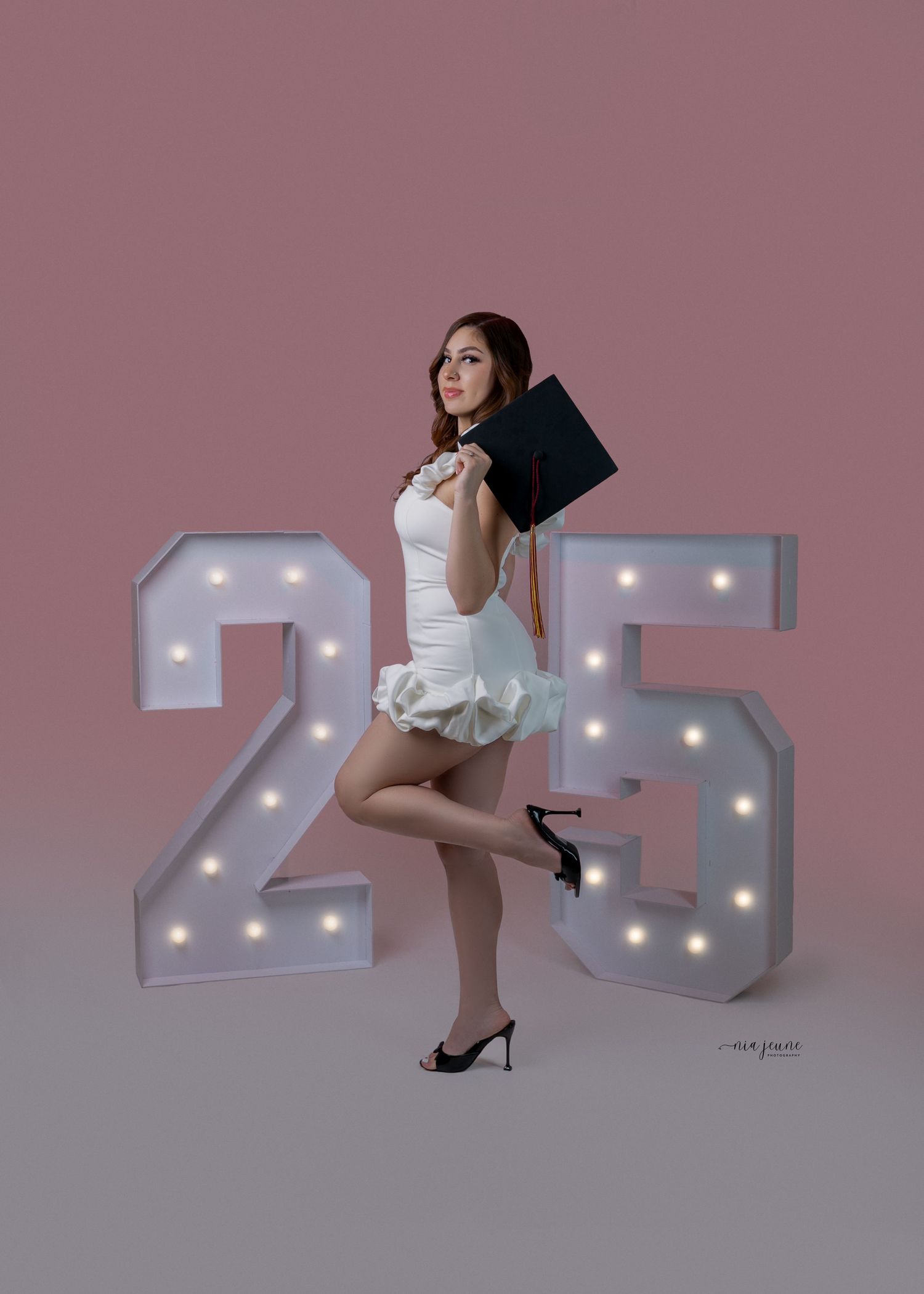 Person in white dress poses elegantly next to illuminated '25' marquee numbers against dusty pink background.