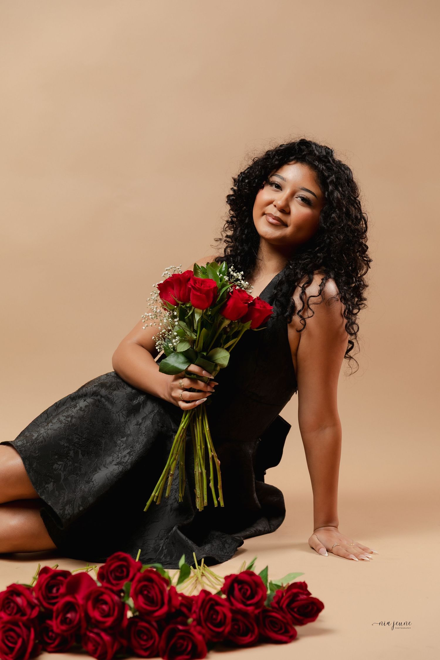 Fashion model in black dress poses elegantly with red roses against beige backdrop in artistic photo series.