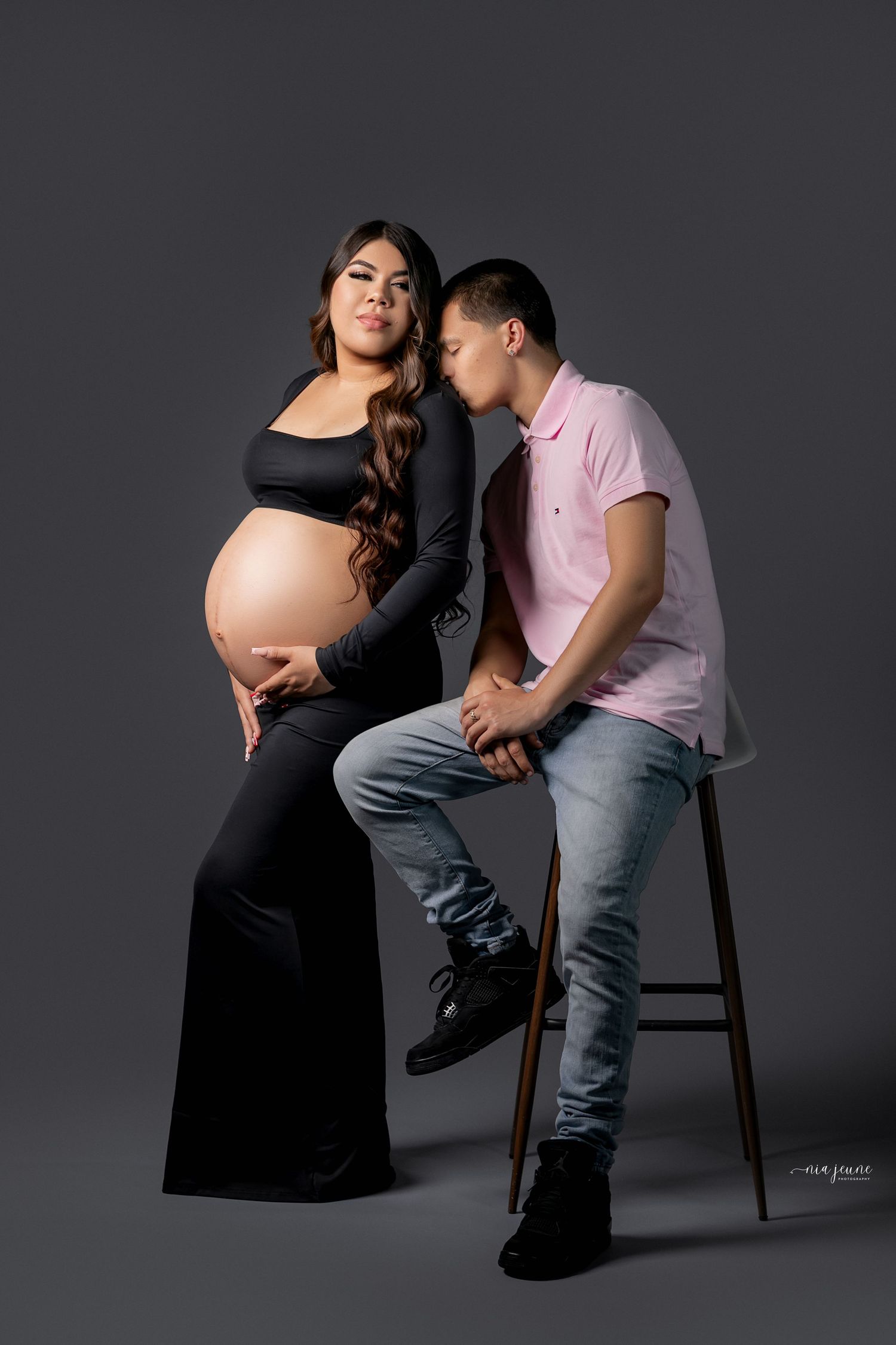 A pregnant woman in a black outfit poses with someone sitting on a stool against a dark studio backdrop.