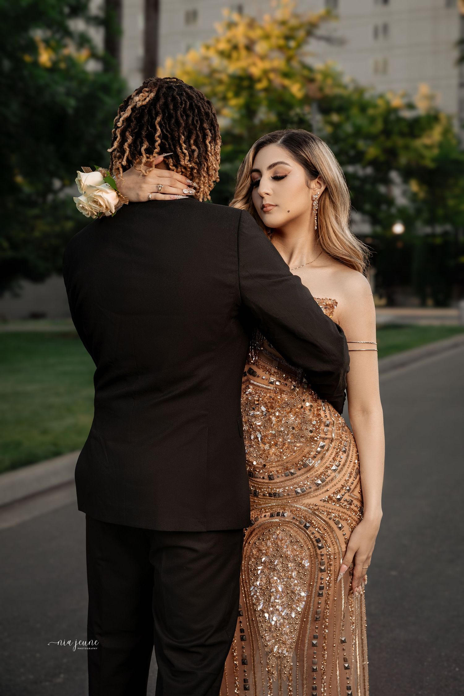 Couple in formal attire sharing intimate moment on path, woman wearing sparkly golden evening gown.
