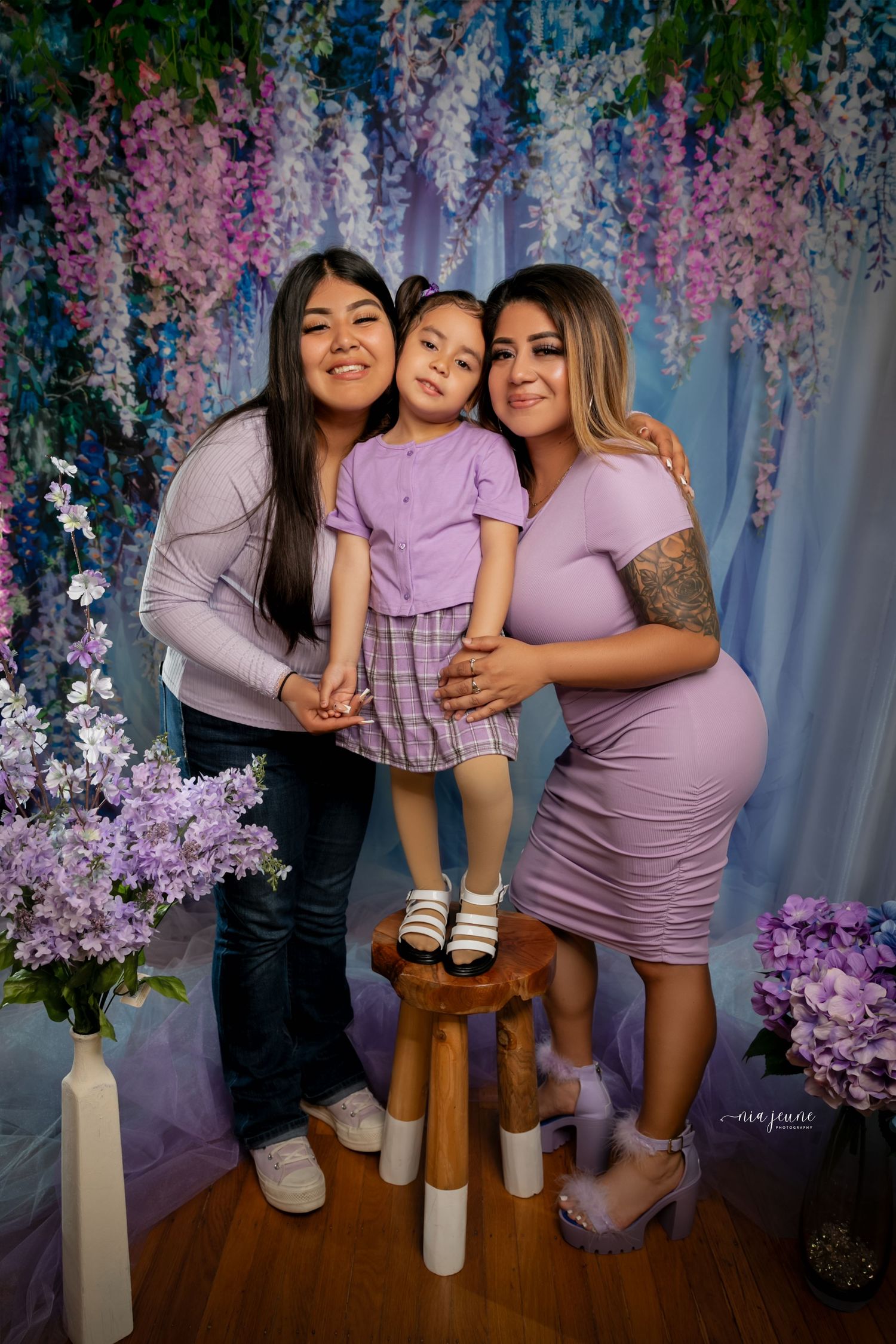 Three generations pose together under hanging purple wisteria flowers in matching lavender outfits.