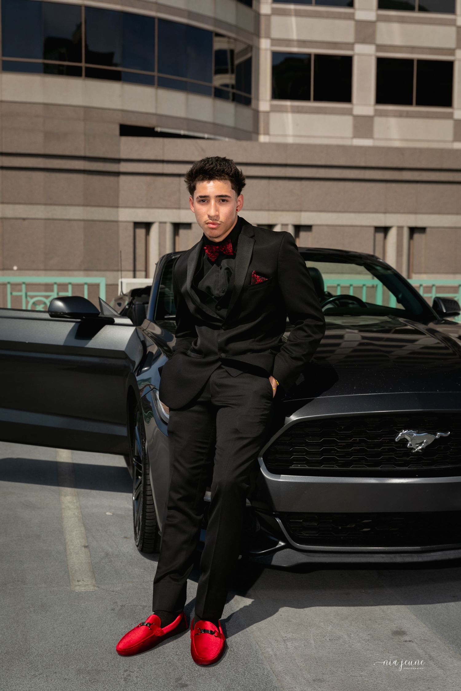 A stylish portrait of someone in all black attire posing with a Ford Mustang in an urban setting.
