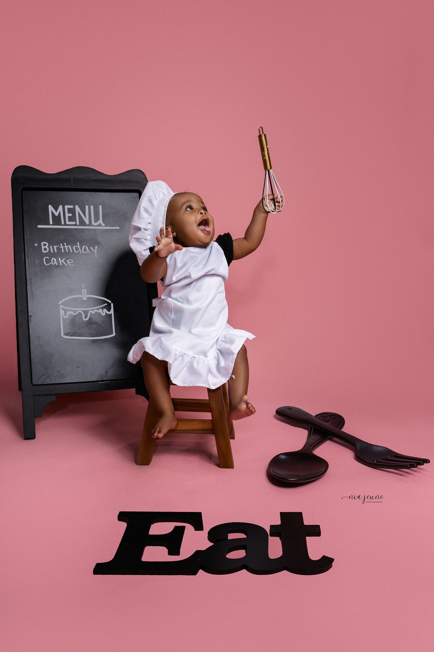Creative baby photo session with menu board props and white dress against pink background.