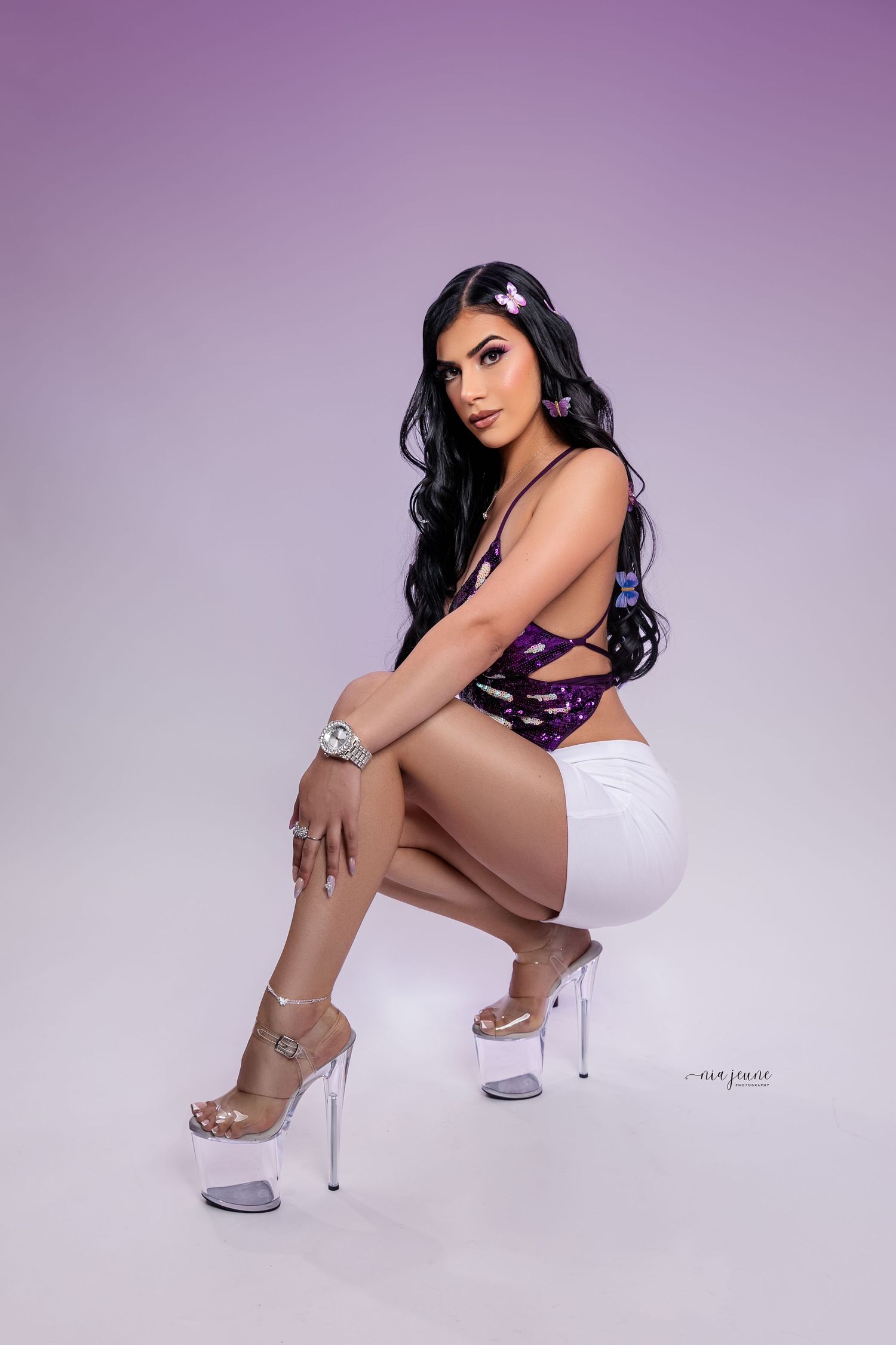 Fashion model in clear platform heels poses in white shorts and purple top against lavender backdrop.