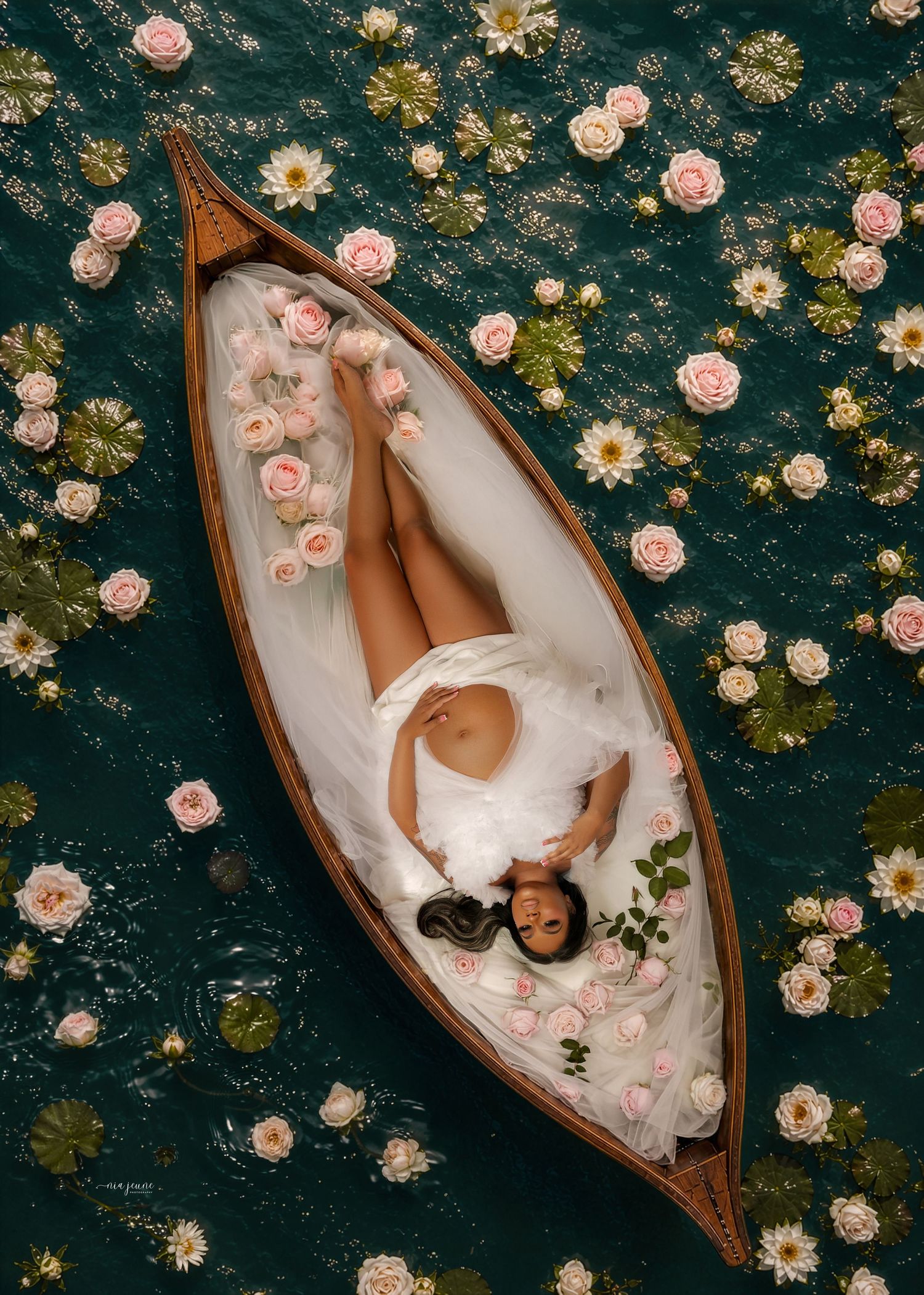Woman in white dress lying in a wooden boat surrounded by pink roses and water lilies on a dark green pond.