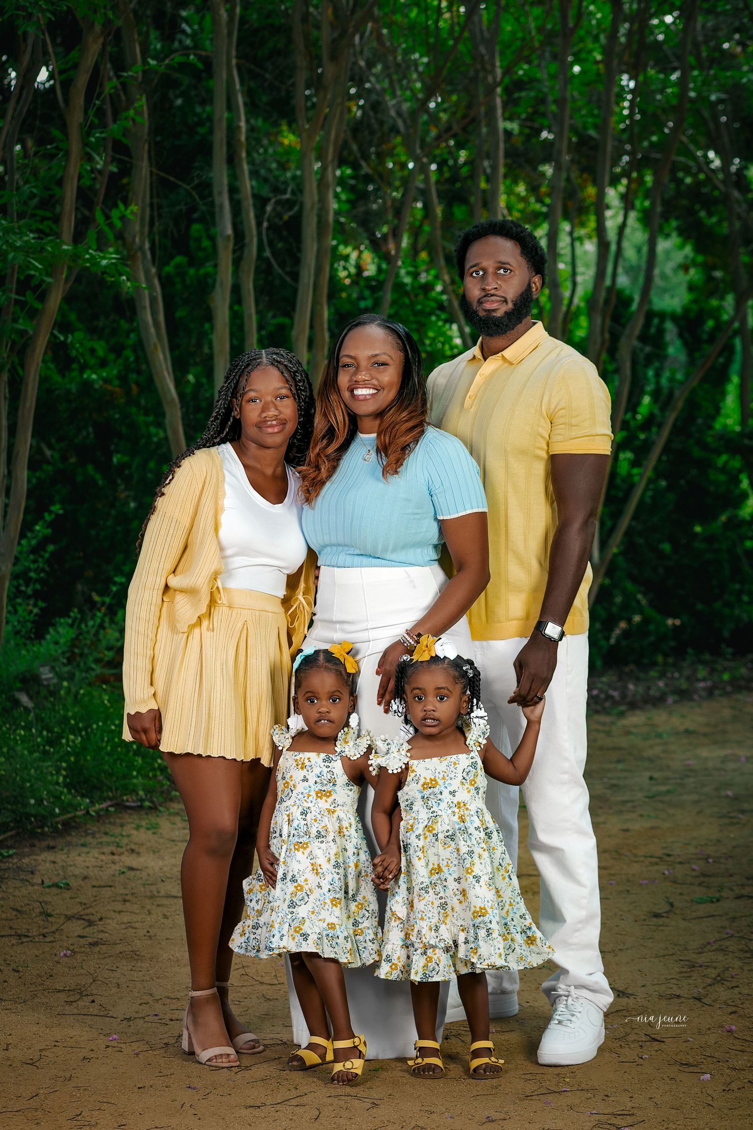 A family of five wearing coordinated yellow, white and floral outfits poses together in a lush outdoor setting.