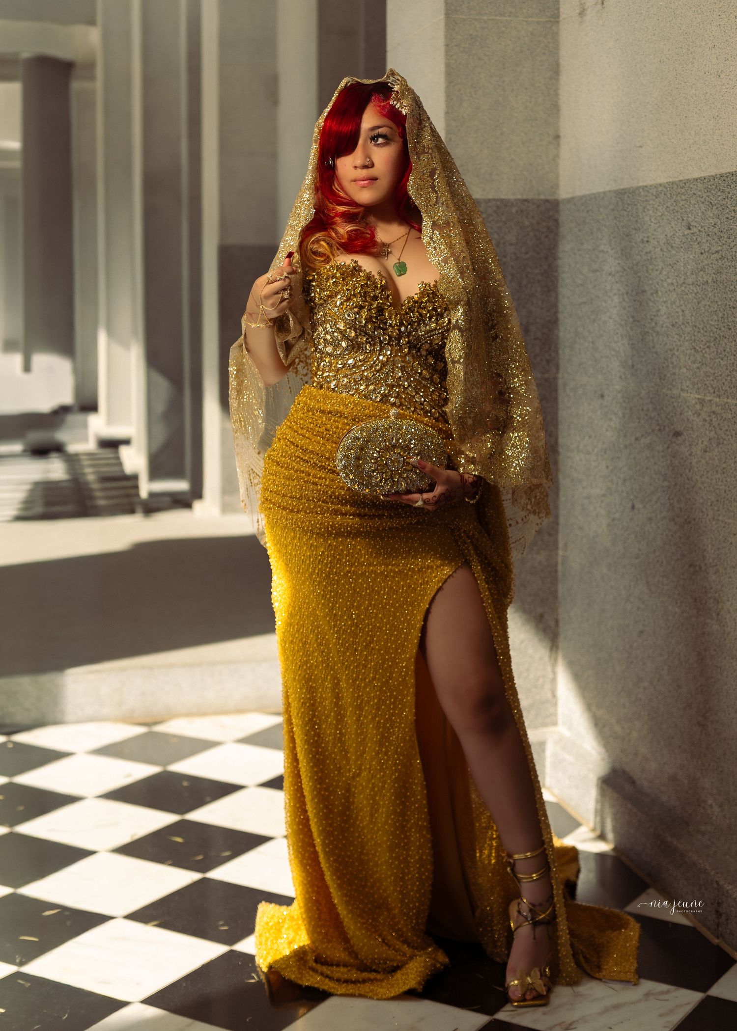Woman in gold sequin gown with veil and red hair poses elegantly on checkered floor by stone columns.