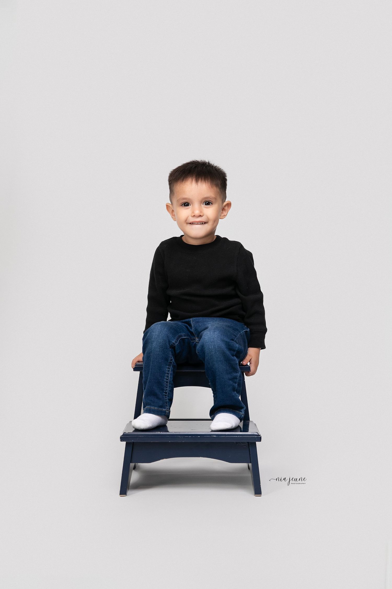 A young boy in black sweater and jeans sits smiling on a small blue stool.