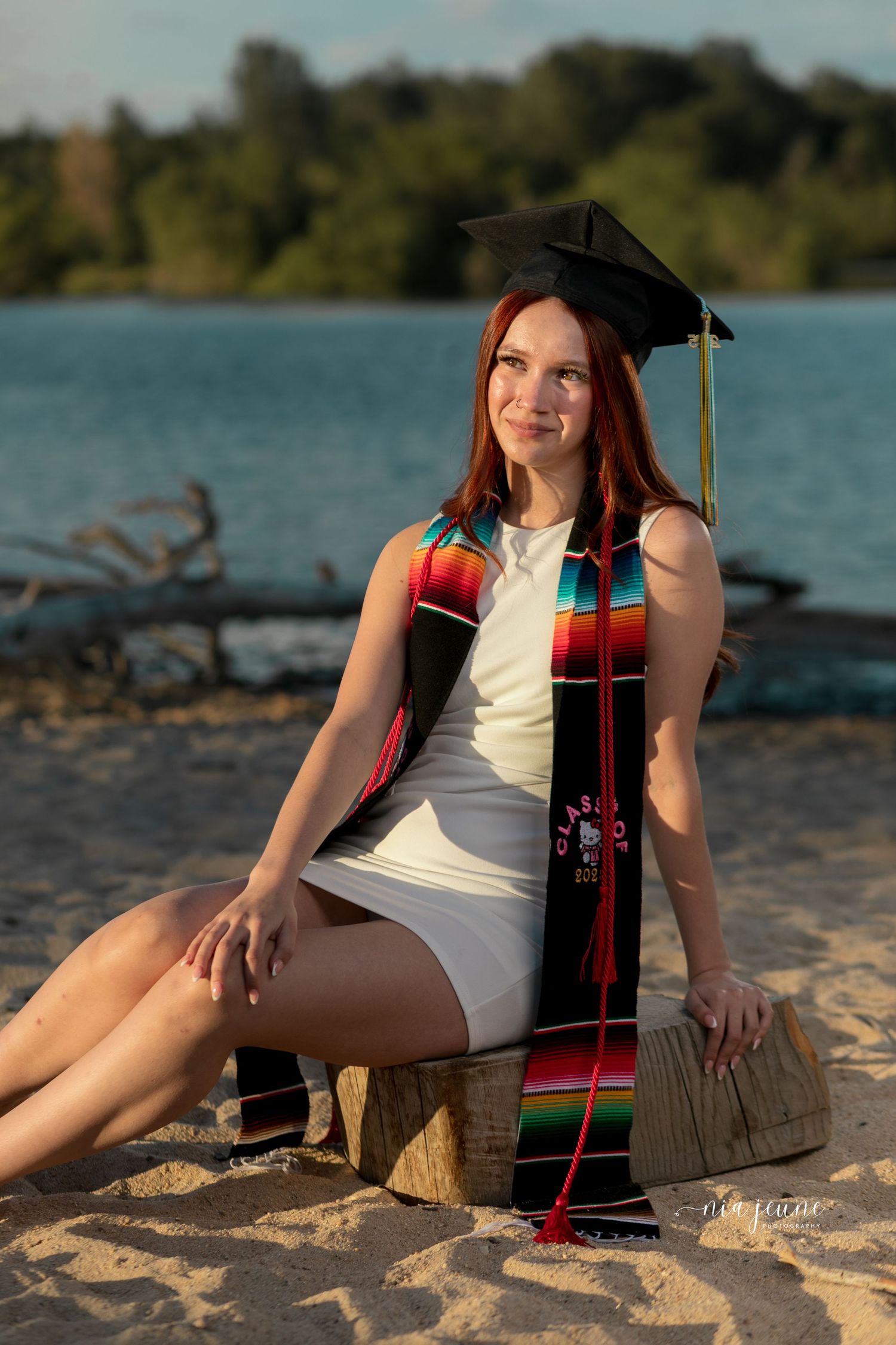 Graduate in white dress and graduation cap poses on beach with colorful stole against lake backdrop.