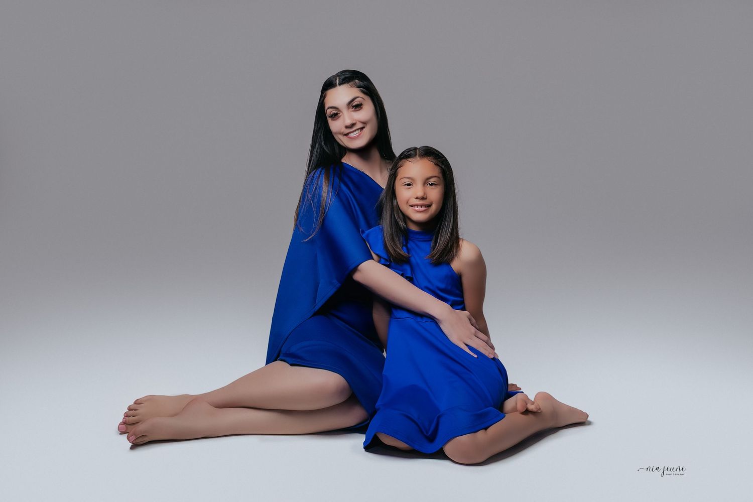 Two people wearing matching royal blue dresses pose together against a gray studio background.