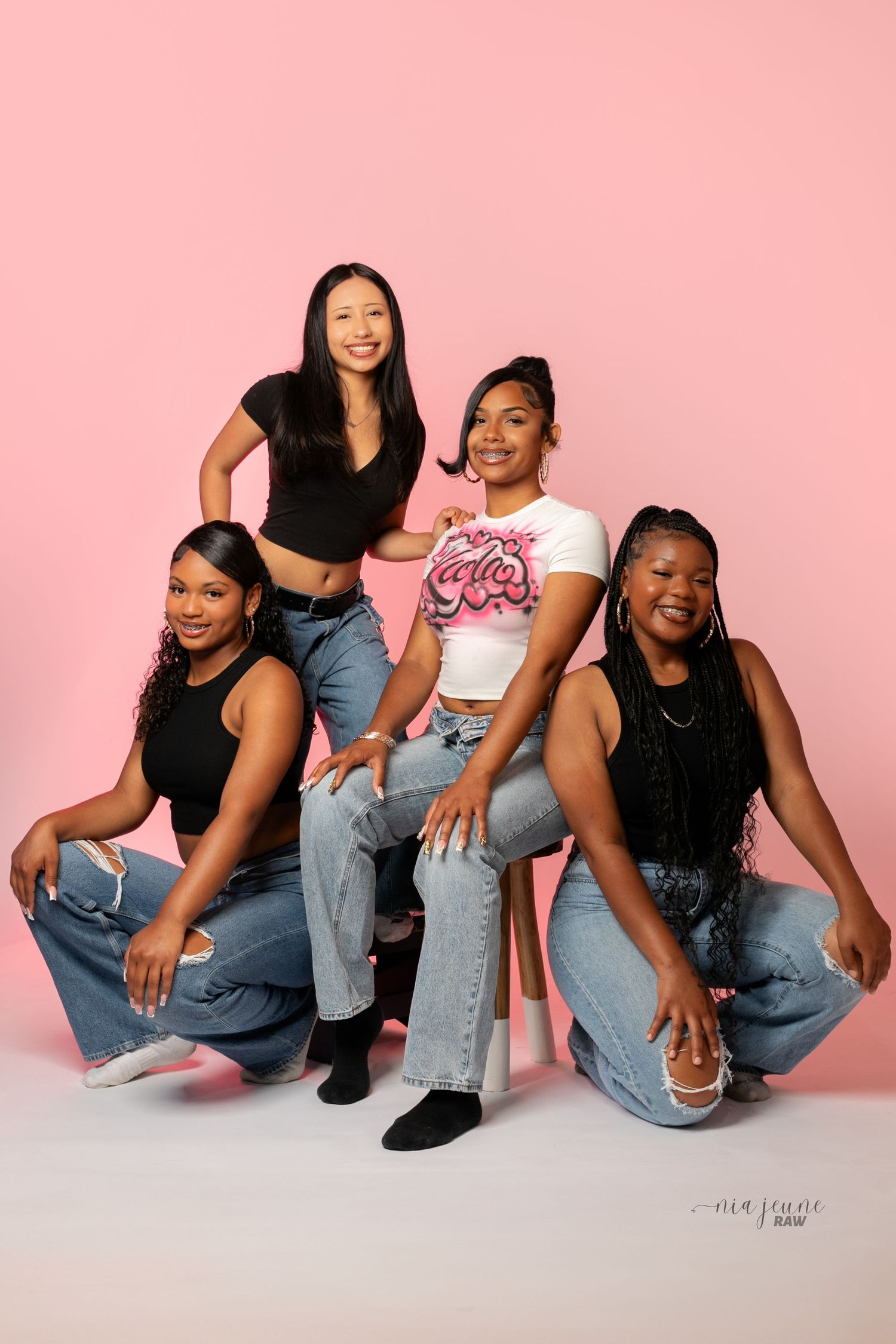Four friends pose together in casual denim outfits against a pink backdrop during a fun photoshoot.